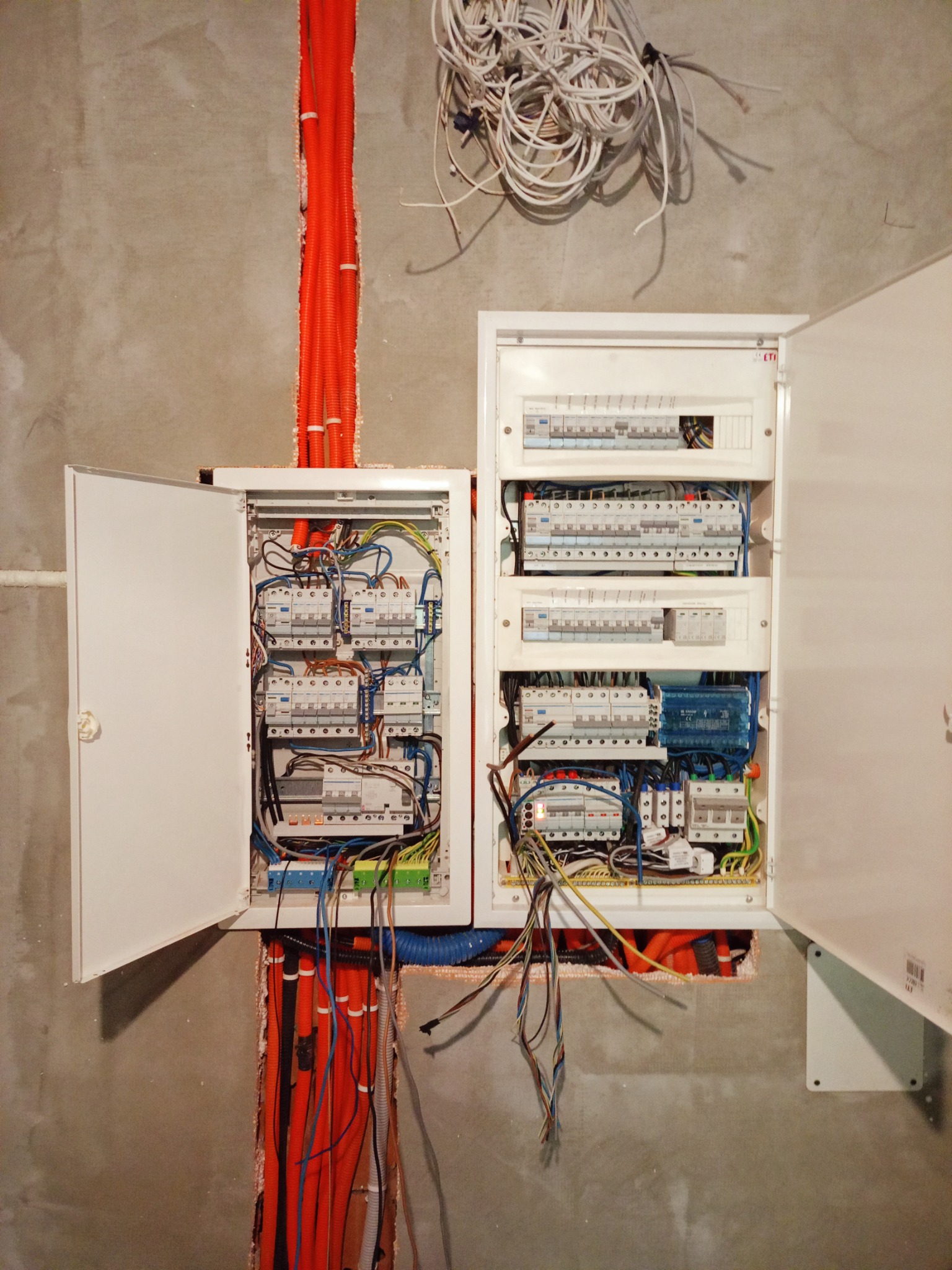 Rozdzielnia elektryczna z otwartymi drzwiczkami, widoczne kable i zabezpieczenia. Pomarańczowe rury instalacyjne w ścianie. Szare tło ściany w surowym stanie.