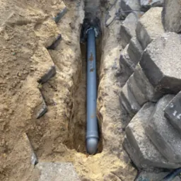 Szary rurociąg kanalizacyjny ułożony w wykopie ziemnym, obok kostka brukowa i piasek, widoczna łopata.