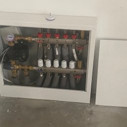 AW BUD - Rozdzielacz ogrzewania podłogowego w szafce, widoczne rury doprowadzające i odprowadzające wodę, manometr oraz numeracja obwodów.