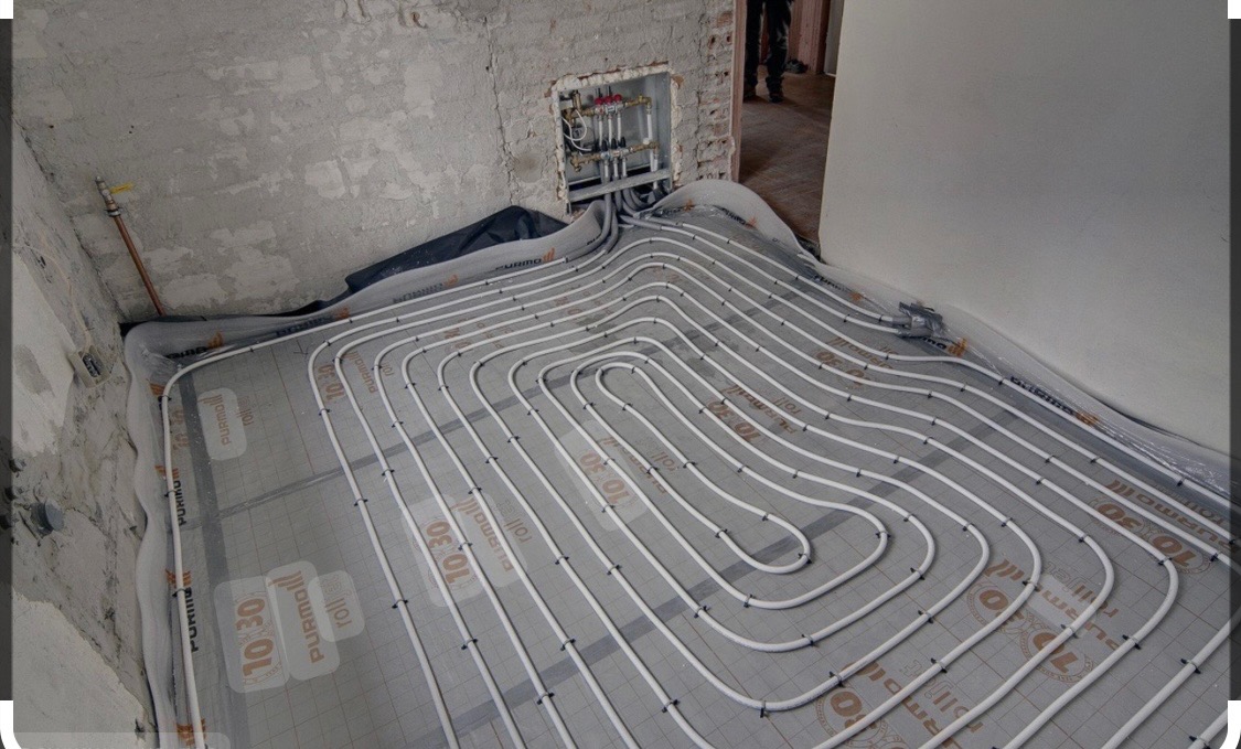 Białe rury systemu grzewczego ułożone na szarej macie izolacyjnej na surowej podłodze w trakcie instalacji, widoczne złącza i kolektor w ścianie.