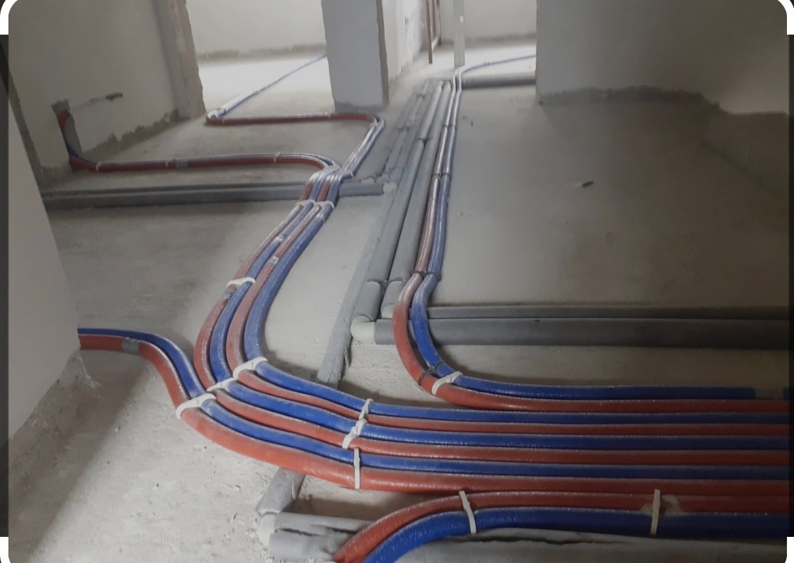 Wiązka rur instalacyjnych, czerwono-niebieskich, prowadzona wzdłuż surowej ściany budynku, z elementami izolacji termicznej, zabezpieczona białymi opaskami zaciskowymi.