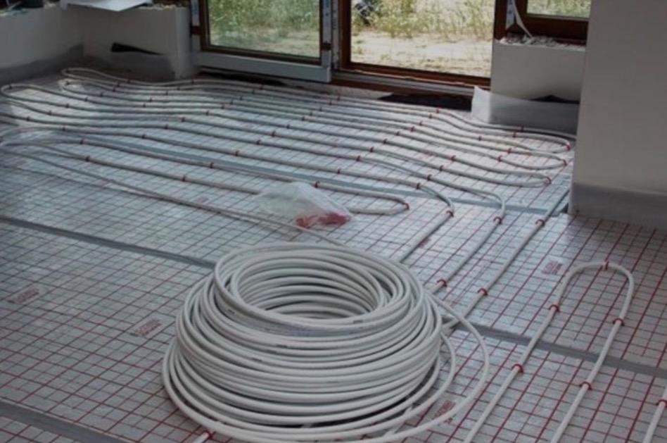 Rozłożone rury ogrzewania podłogowego na siatce z rolką niezamontowanych rur w pomieszczeniu z oknem.