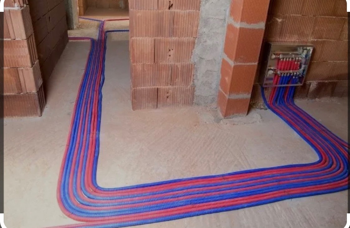 Instalacja ogrzewania podłogowego w surowym stanie, widoczne rury grzewcze ułożone na posadzce, rozdzielacz zamontowany na ścianie z cegły.