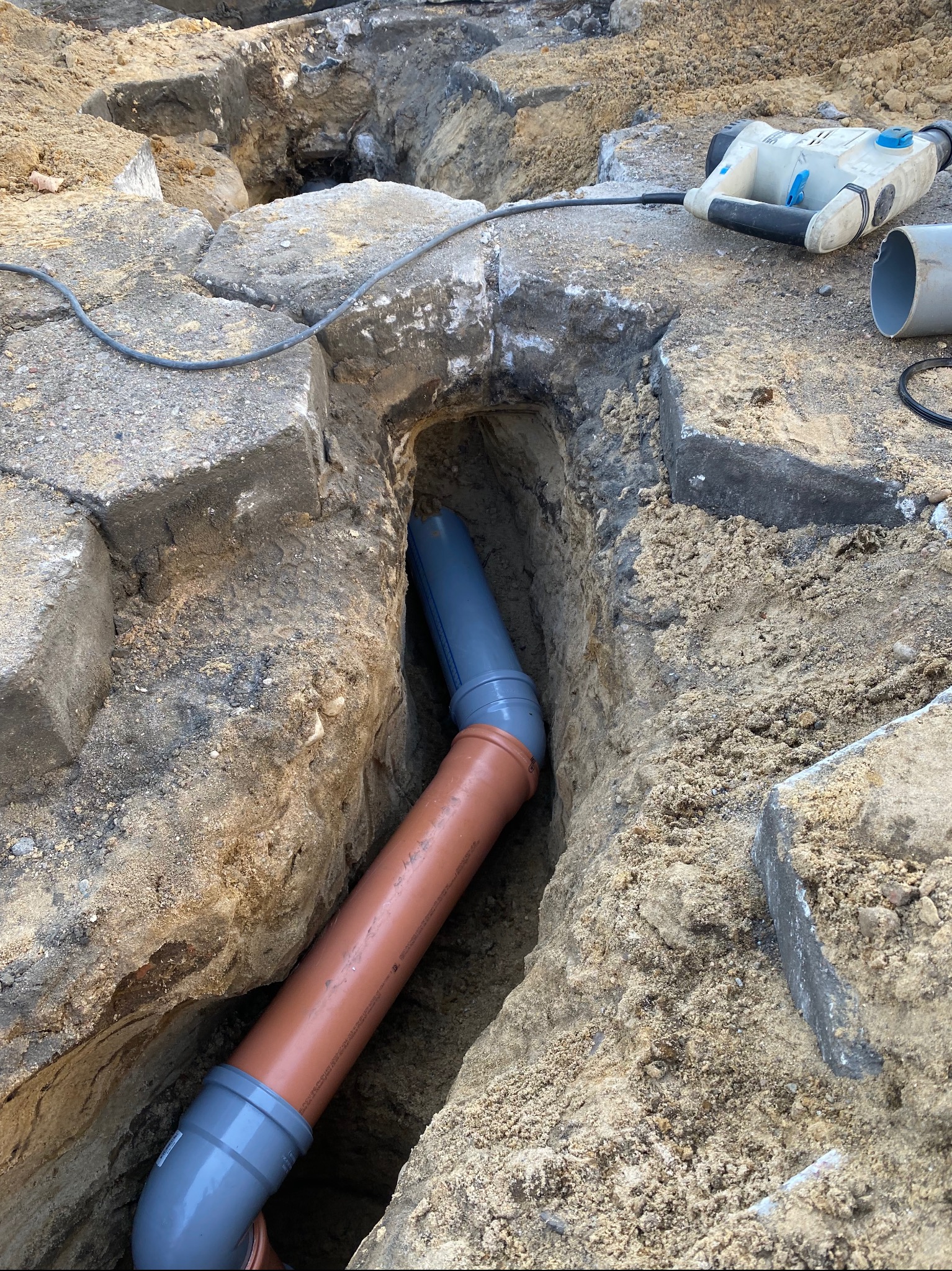 Wykop z ułożonymi rurami kanalizacyjnymi z tworzywa sztucznego, widoczny młot udarowy i fragmenty gruzu na powierzchni.