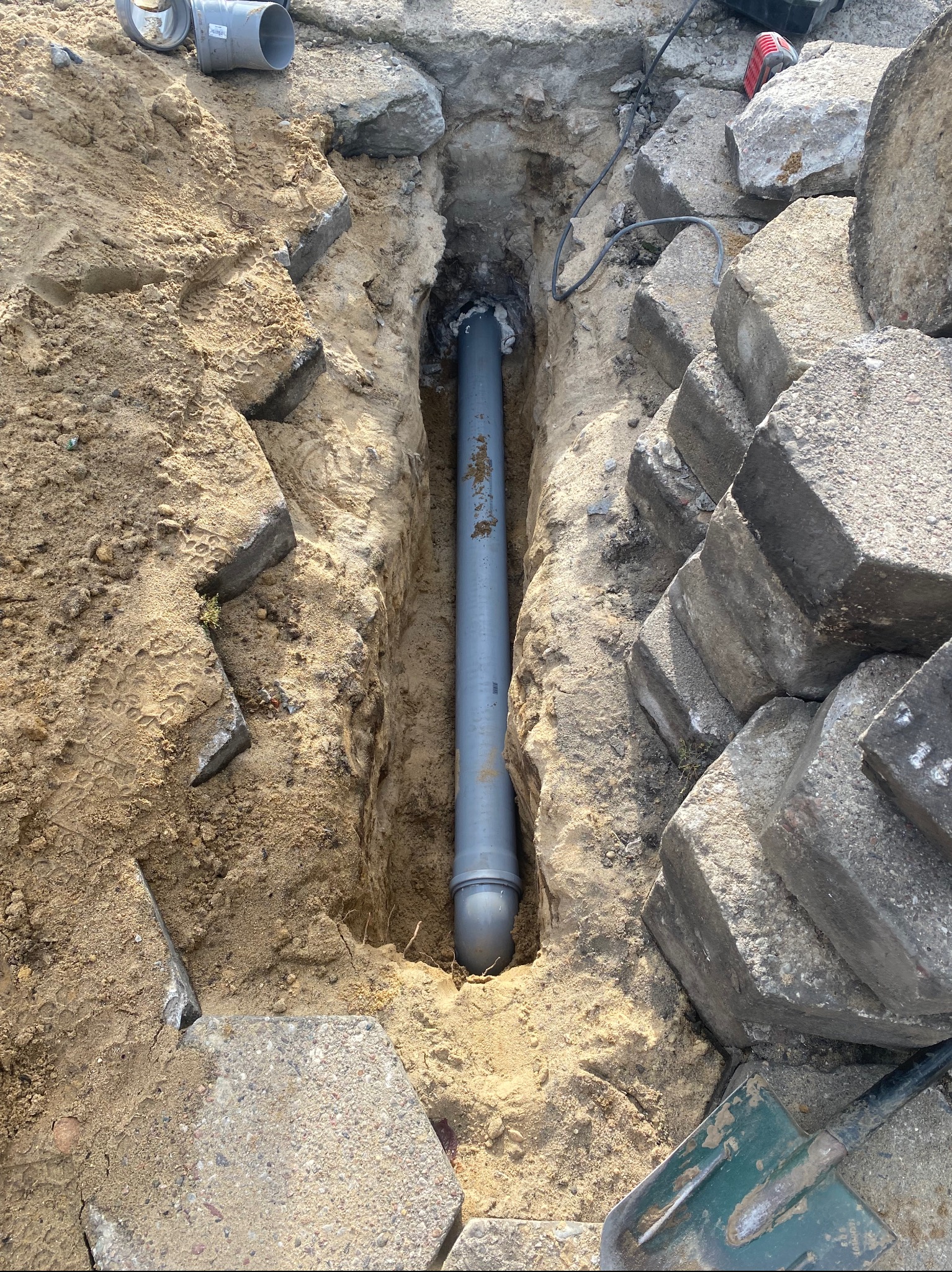 Szary rurociąg kanalizacyjny ułożony w wykopie ziemnym, obok kostka brukowa i piasek, widoczna łopata.