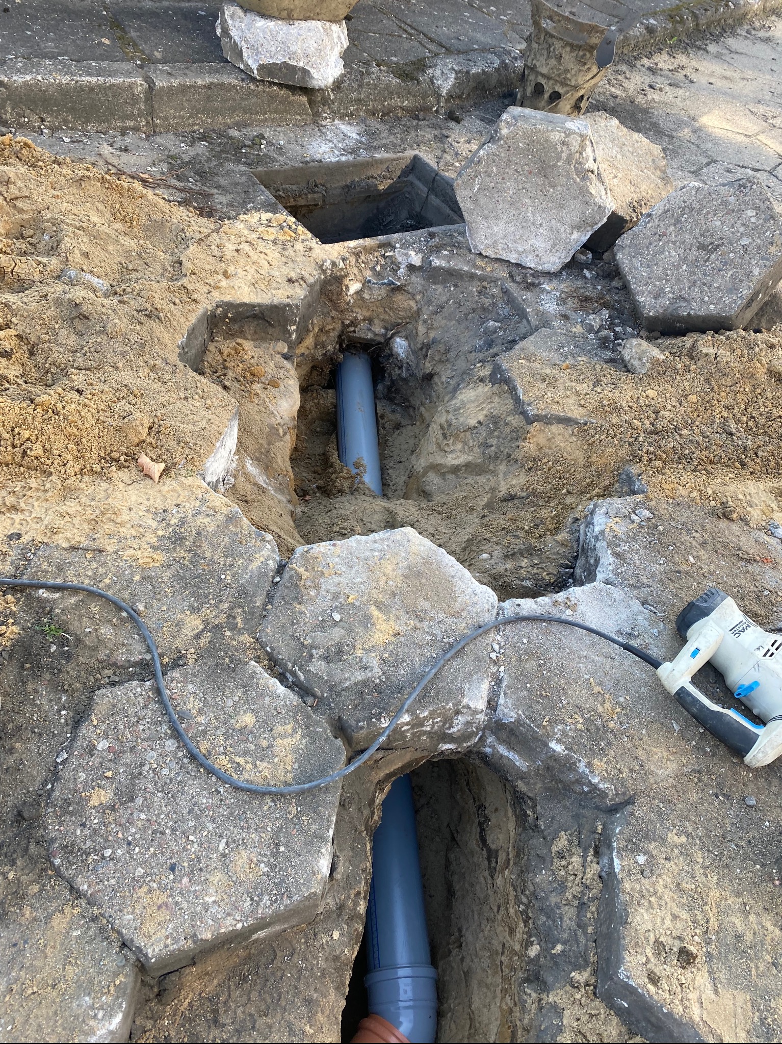 Wykop z widocznymi fragmentami rur kanalizacyjnych, otoczony gruzem i ziemią, z elektrycznym młotem udarowym leżącym obok.