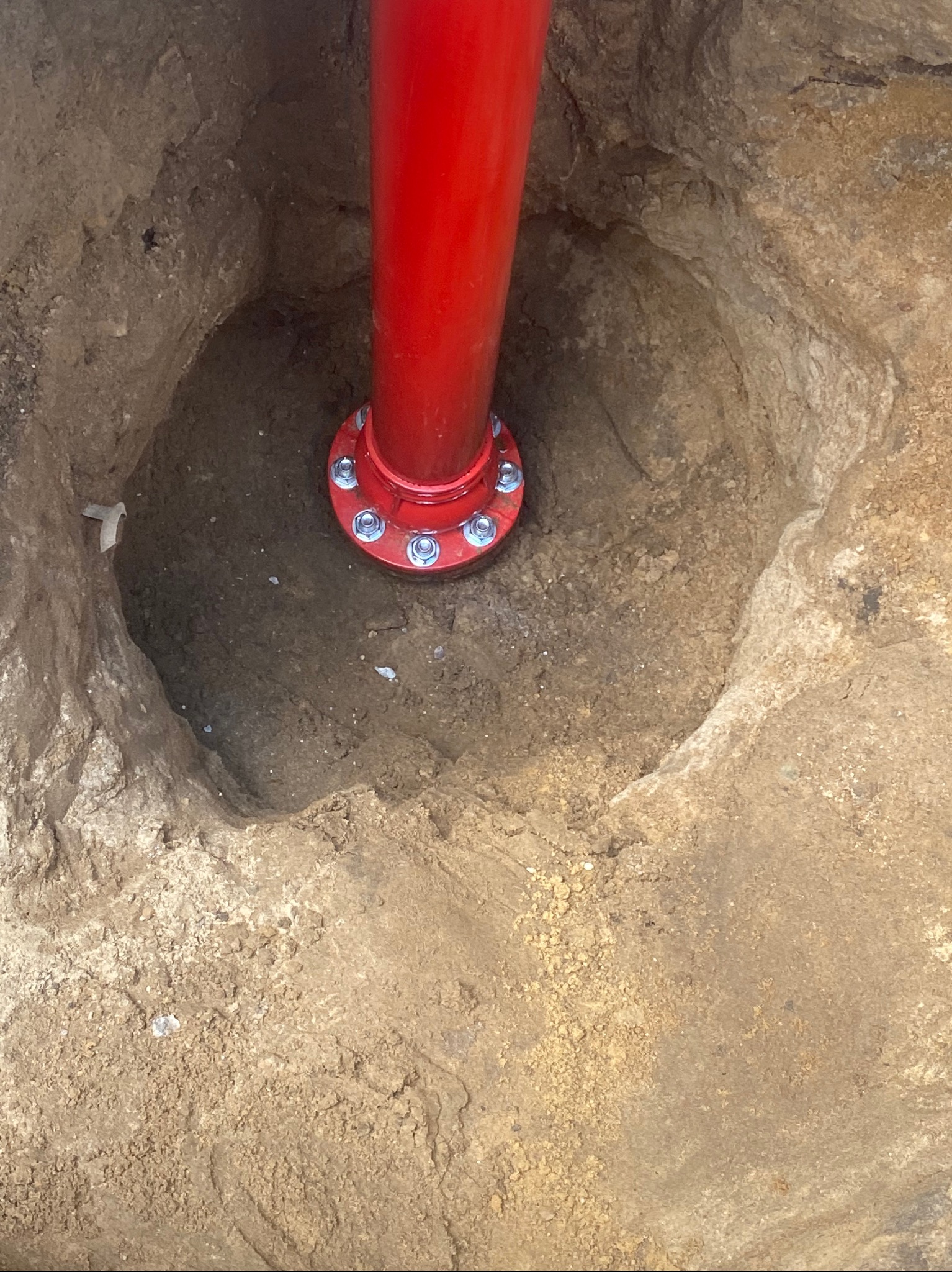 Czerwona rura z kołnierzem i śrubami, wystająca z wykopu w ziemi. Widoczny fragment papieru na ścianie wykopu.