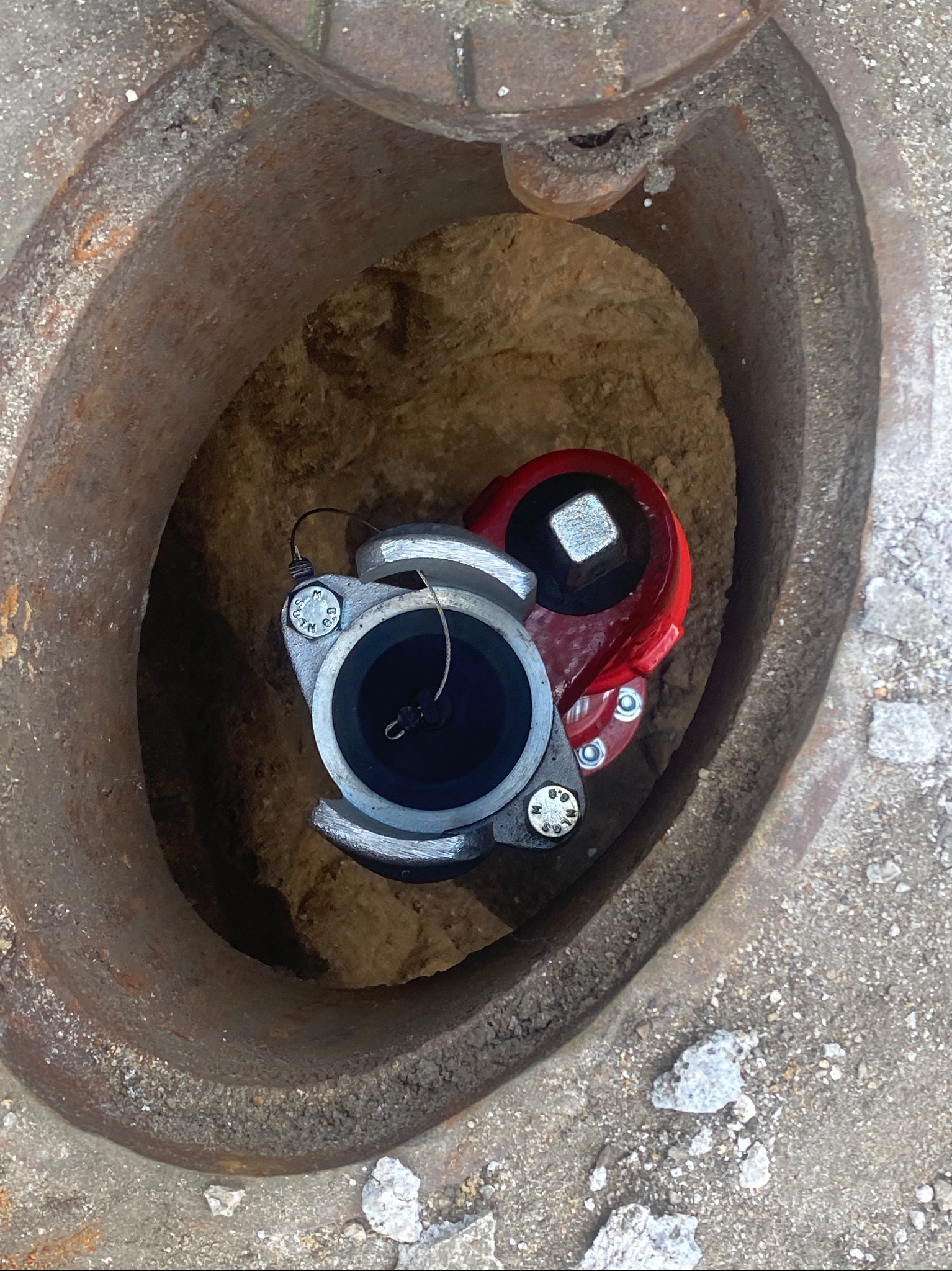 Nowe przyłącze wodociągowe z czerwoną zasuwa zainstalowane w studzience kanalizacyjnej z betonowym obrzeżem.