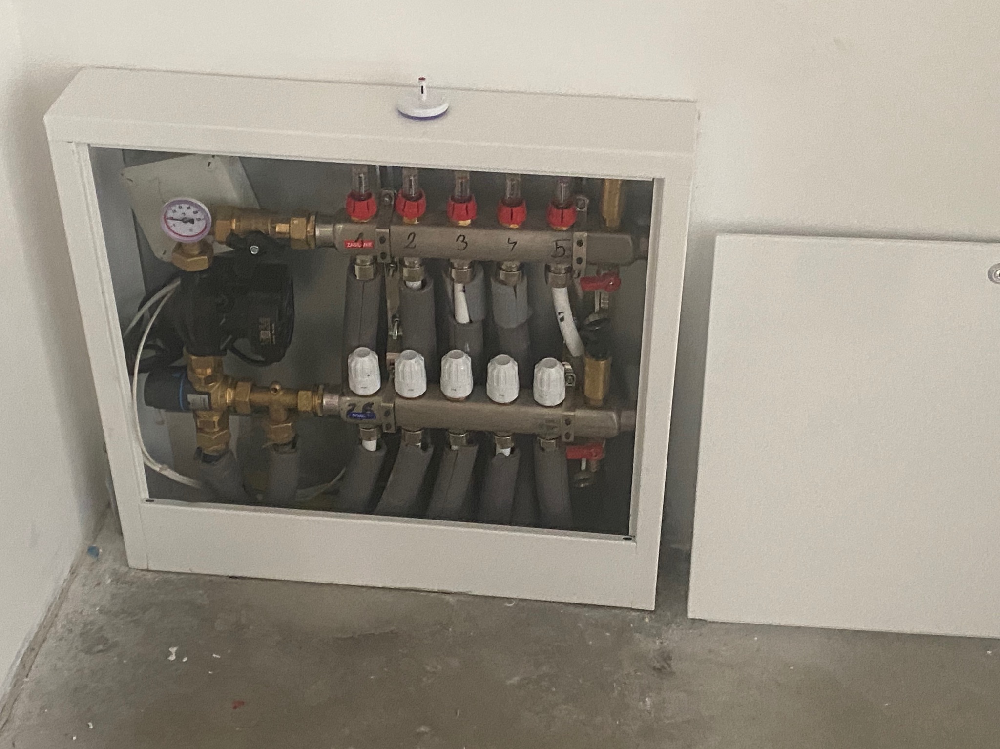Rozdzielacz ogrzewania podłogowego w szafce, widoczne rury doprowadzające i odprowadzające wodę, manometr oraz numeracja obwodów.
