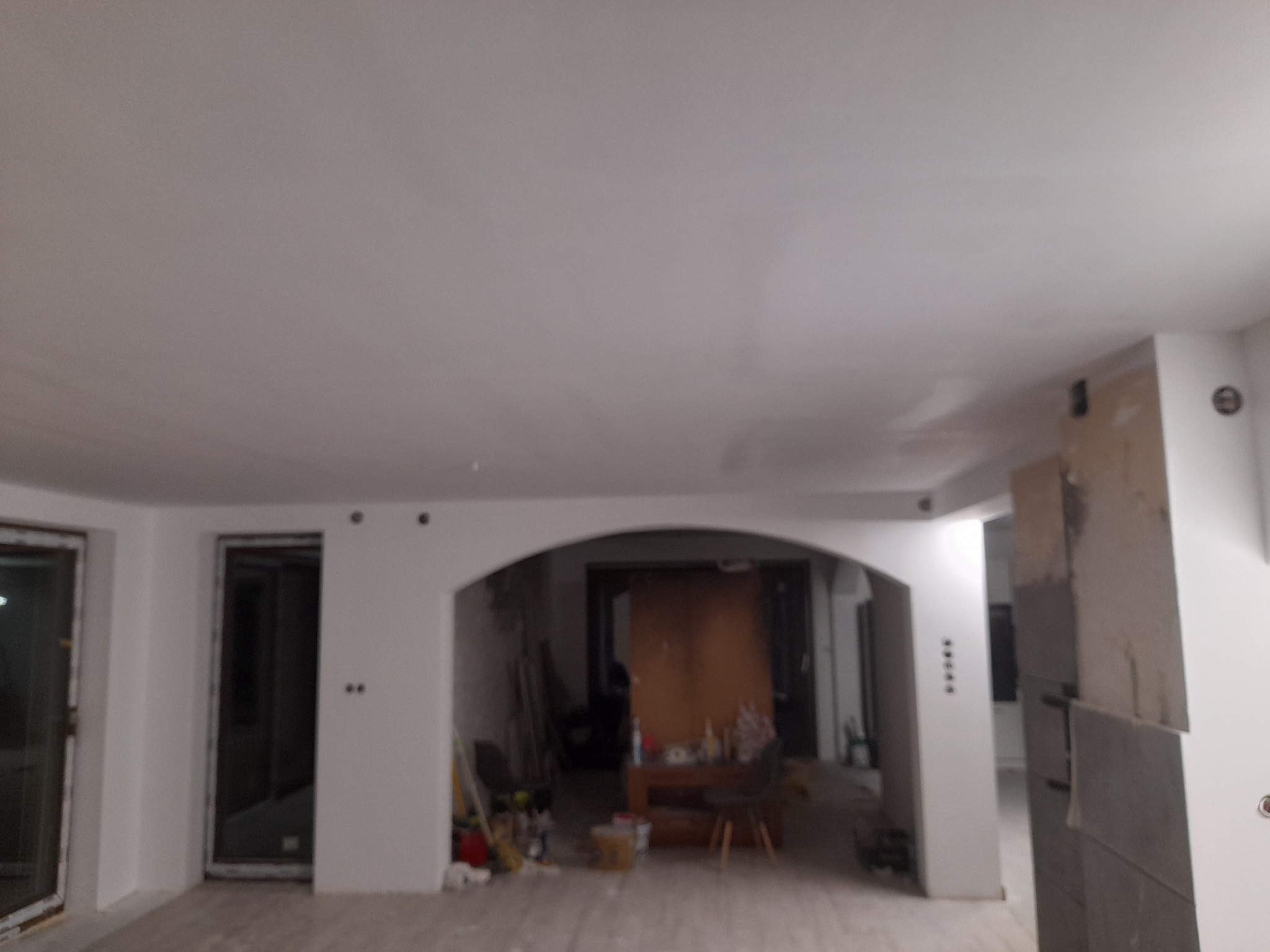 Wnętrze w trakcie remontu, widoczne świeżo wygładzone ściany i sufit, łukowate przejście, częściowo zakryte meble i narzędzia malarskie w tle, na filarze widoczne ślady po szpachlowaniu.