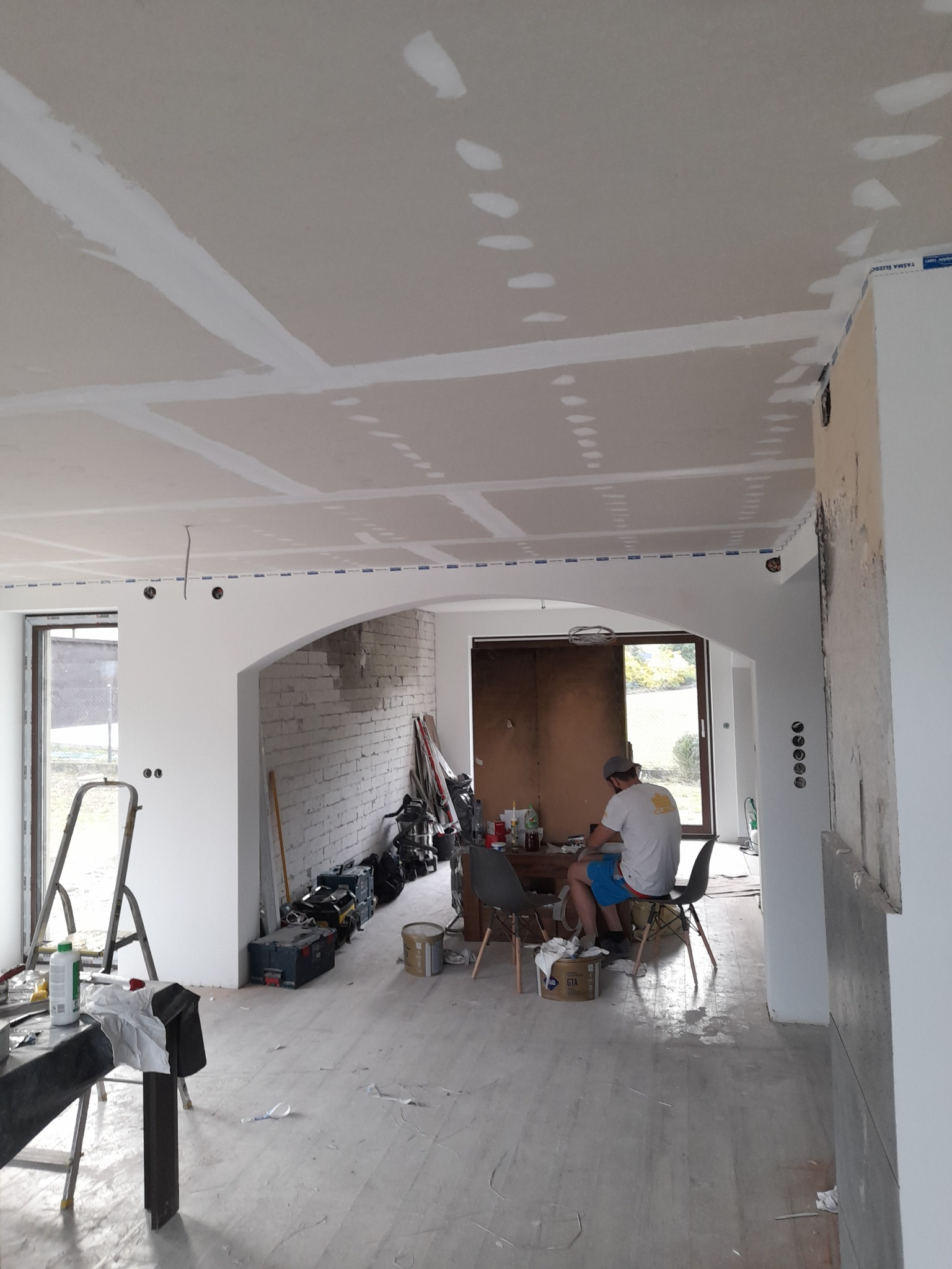 Wnętrze w trakcie remontu: widoczne łączenia płyt kartonowo-gipsowych na suficie i ścianach wypełnione masą szpachlową, narzędzia i materiały budowlane porozrzucane na podłodze, mężczyzna siedzący...