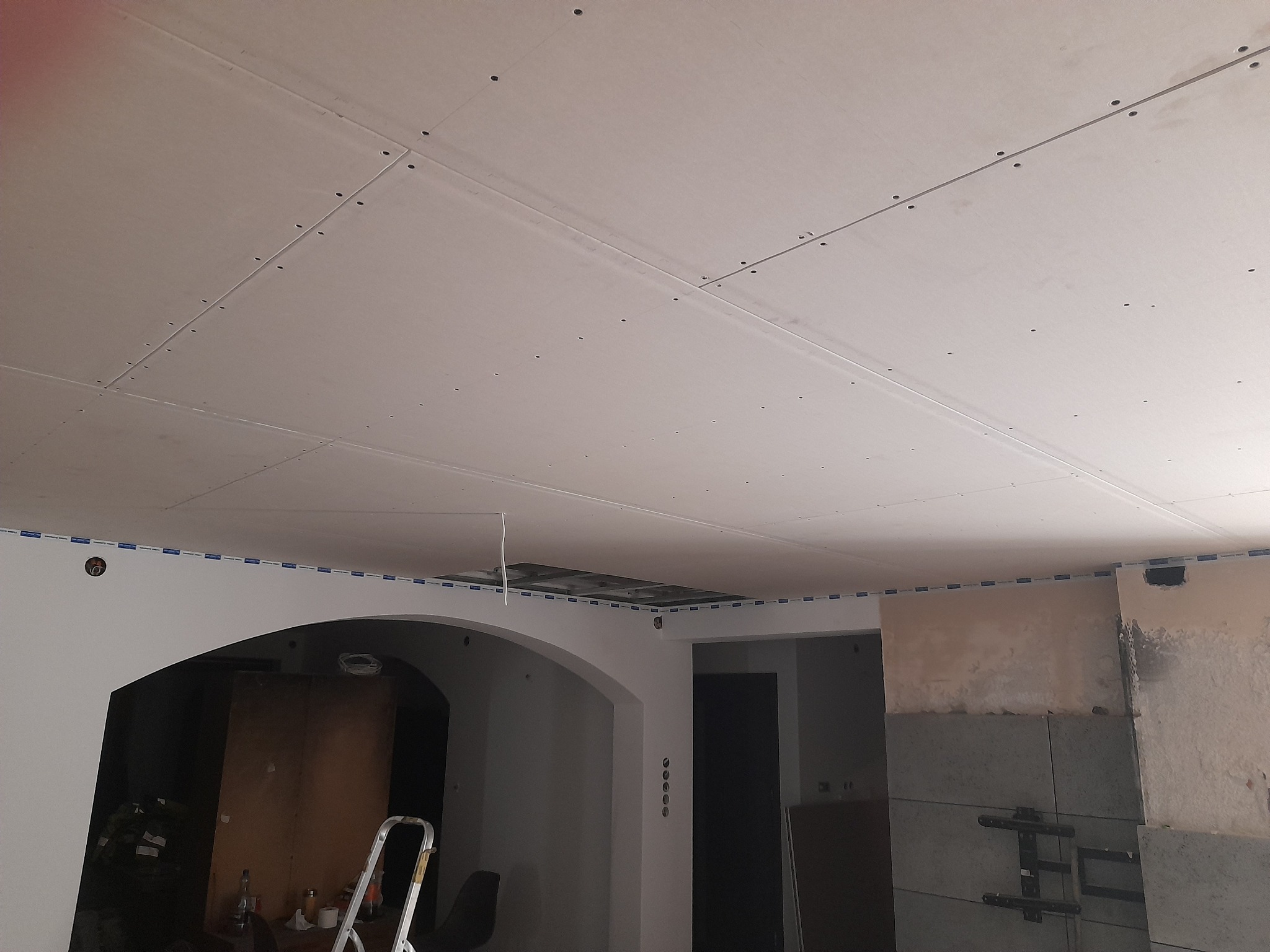 Montaż płyt kartonowo-gipsowych na suficie, widoczne łączenia płyt i śruby mocujące, fragment ściany z widocznym wykończeniem i przygotowaniem do dalszych prac.