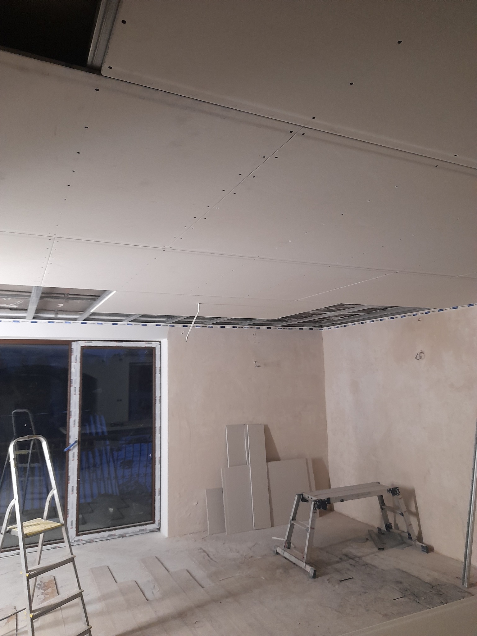 Wnętrze w trakcie montażu sufitu z płyt kartonowo-gipsowych na metalowej konstrukcji, widoczne płyty opierające się o ścianę, drabina aluminiowa i kozioł roboczy.
