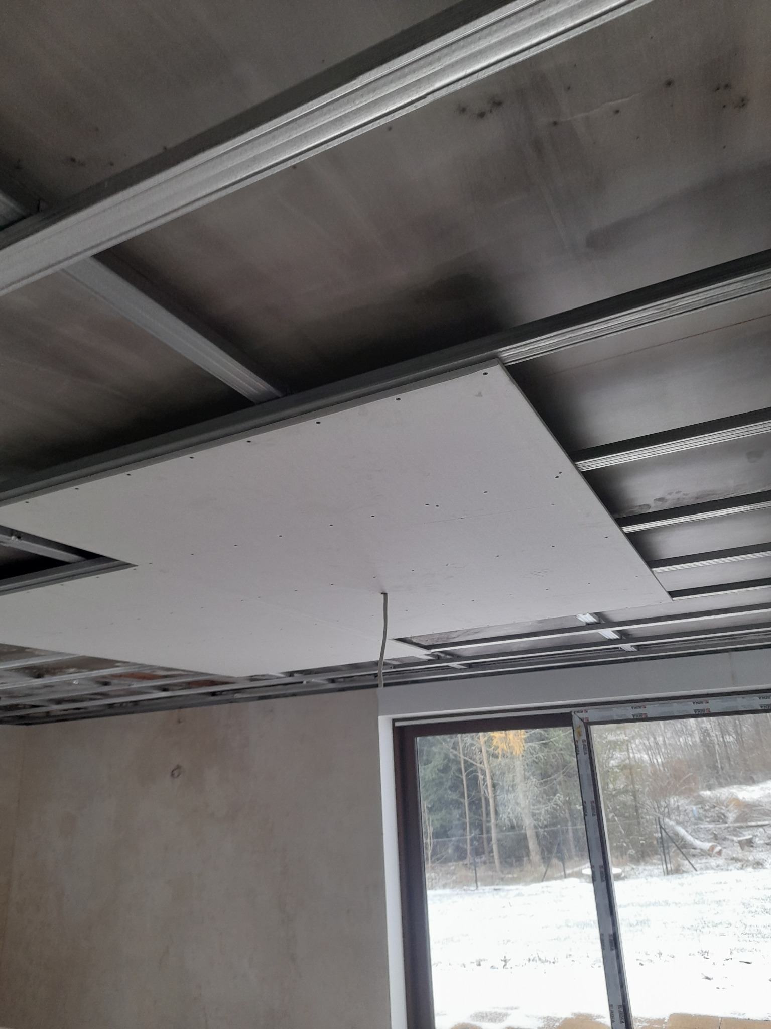 Montaż sufitu podwieszanego z płyt karton-gips na metalowej konstrukcji szkieletowej, widok od dołu, z widocznym oknem i zimowym krajobrazem na zewnątrz.
