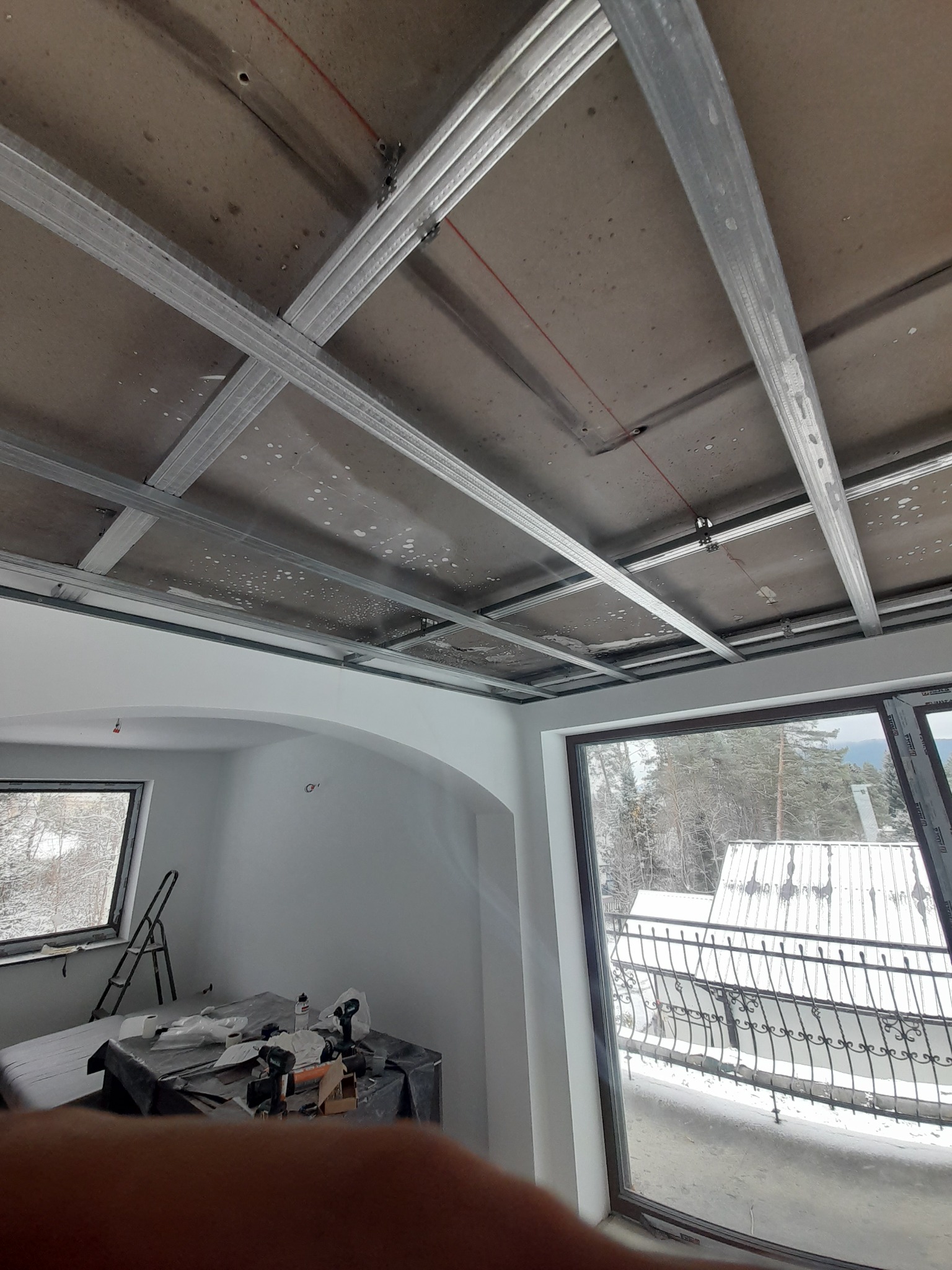 Stelaż z metalowych profili pod montaż sufitu podwieszanego z płyty karton gips, widok z dołu, w tle okno z widokiem na zimowy krajobraz.