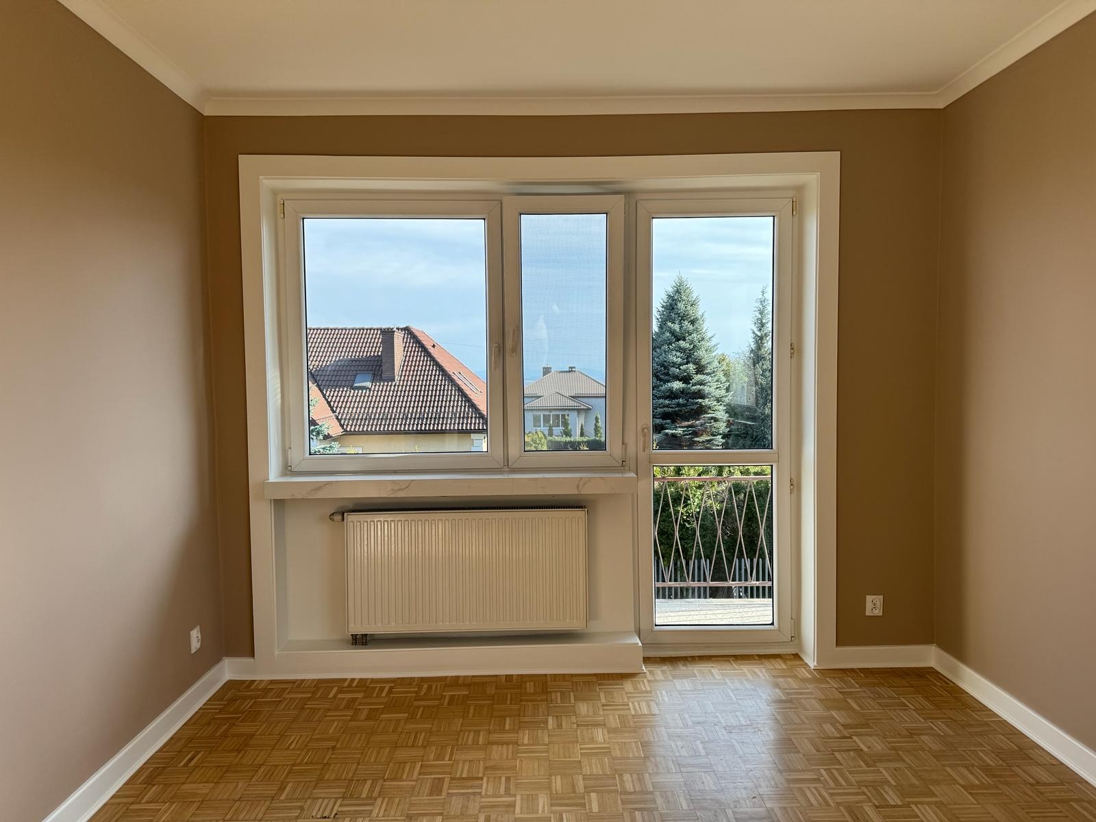 Pokój po odświeżeniu z pomalowanymi ścianami na kolor beżowy, parkietem w jodełkę i oknem z widokiem na domy i drzewa.