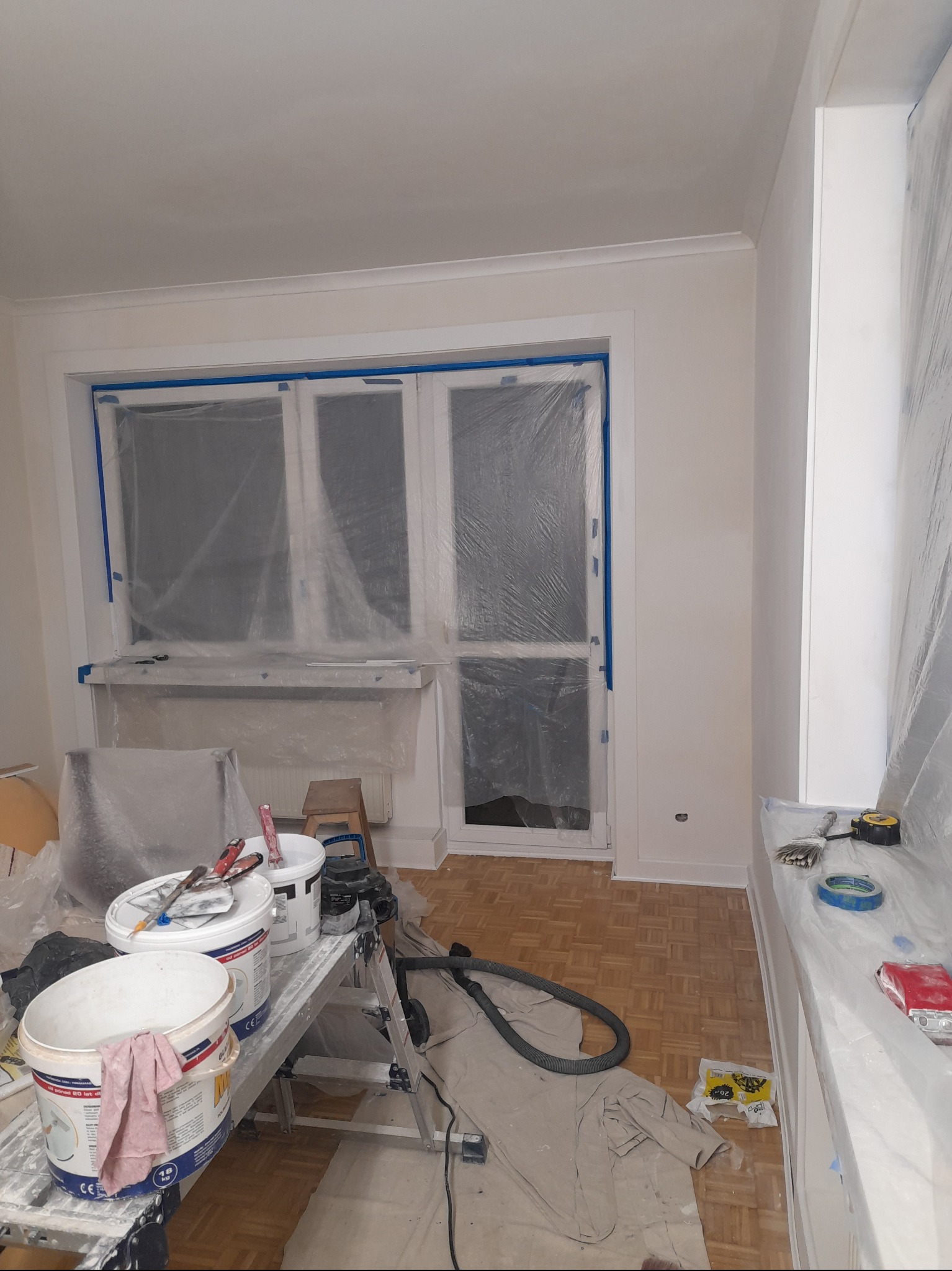Pokój w trakcie prac malarskich: okno zabezpieczone folią i taśmą, wiadra z farbą na drabince, widoczny parkiet na podłodze, ściany przygotowane do malowania.