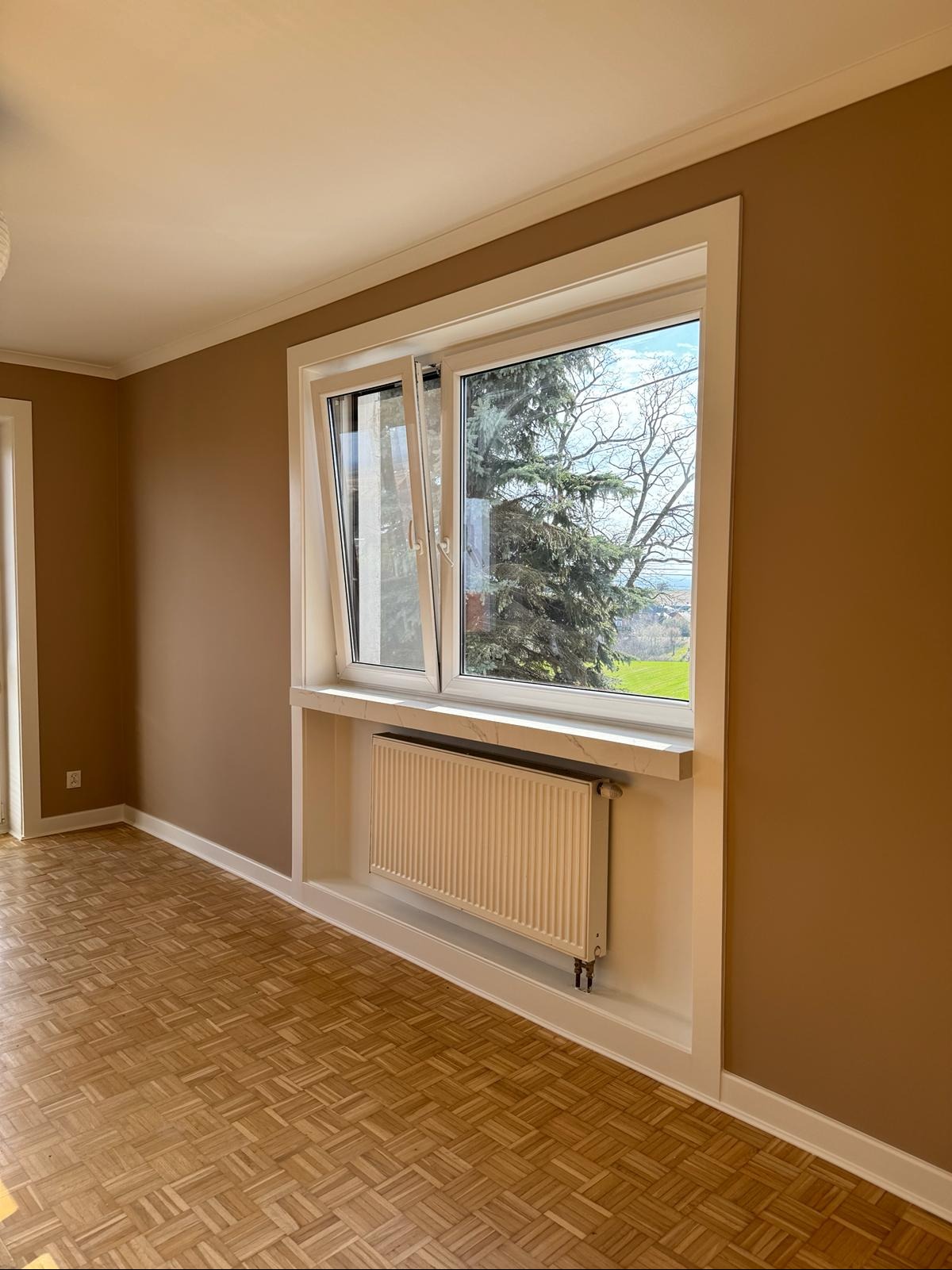 Jasnobrązowe ściany w pokoju z parkietem, biała rama okienna z widokiem na zieleń, biały grzejnik w zabudowie pod oknem.