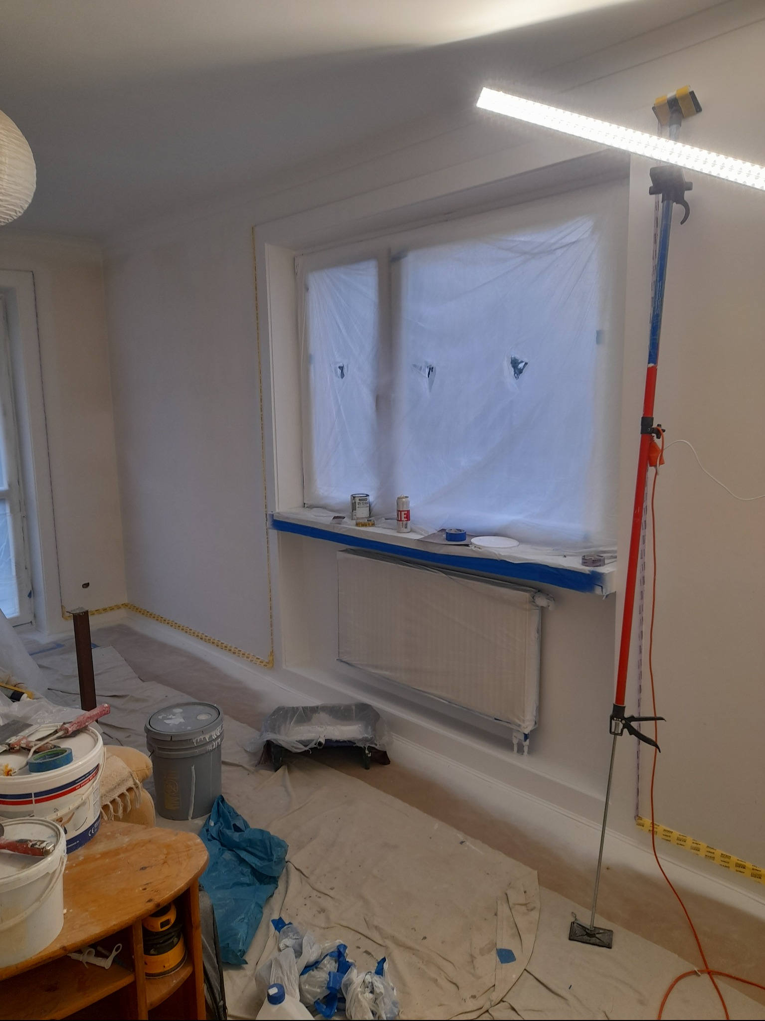 Pokój w trakcie zabezpieczania przed malowaniem: okna zakryte folią, grzejnik i listwy przypodłogowe zabezpieczone taśmą malarską, podłoga przykryta folią ochronną, widoczne wiadra z farbą...