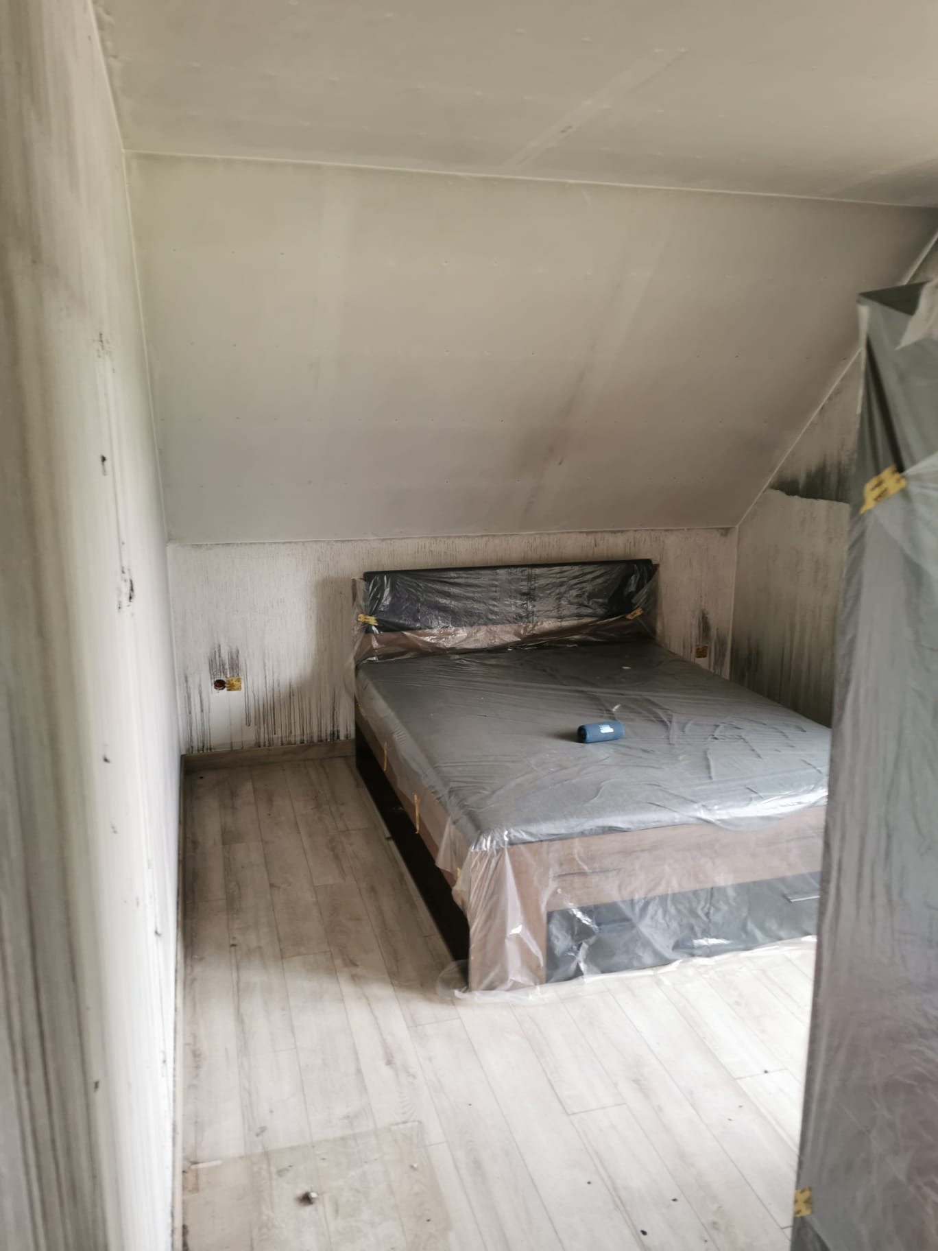 Pokój w trakcie przygotowania do prac wykończeniowych: ściany ze śladami zacieków, podłoga częściowo zabezpieczona, łóżko przykryte folią ochronną, widoczne gniazdko elektryczne.