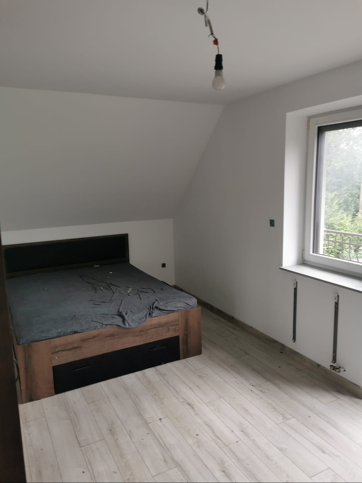 Pokój na poddaszu ze skośnym sufitem, świeżo pomalowany na biało, z drewnianą podłogą i łóżkiem z szarą pościelą, okno z widokiem na zieleń.