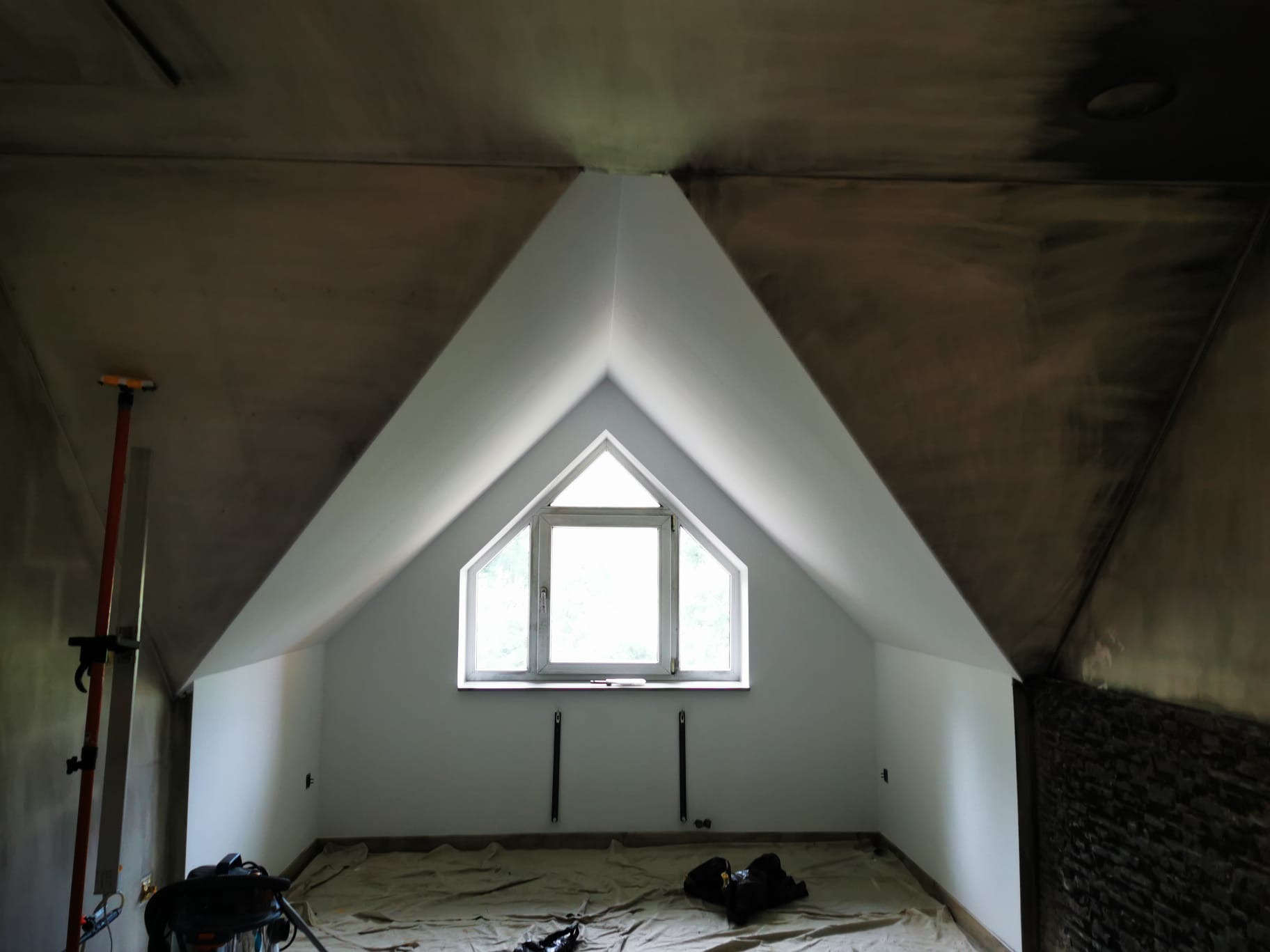 Poddasze w trakcie prac wykończeniowych: pomalowane ściany z oknem w szczycie, sufit częściowo w stanie surowym, podłoga zabezpieczona folią malarską, widoczne narzędzia i drabina.