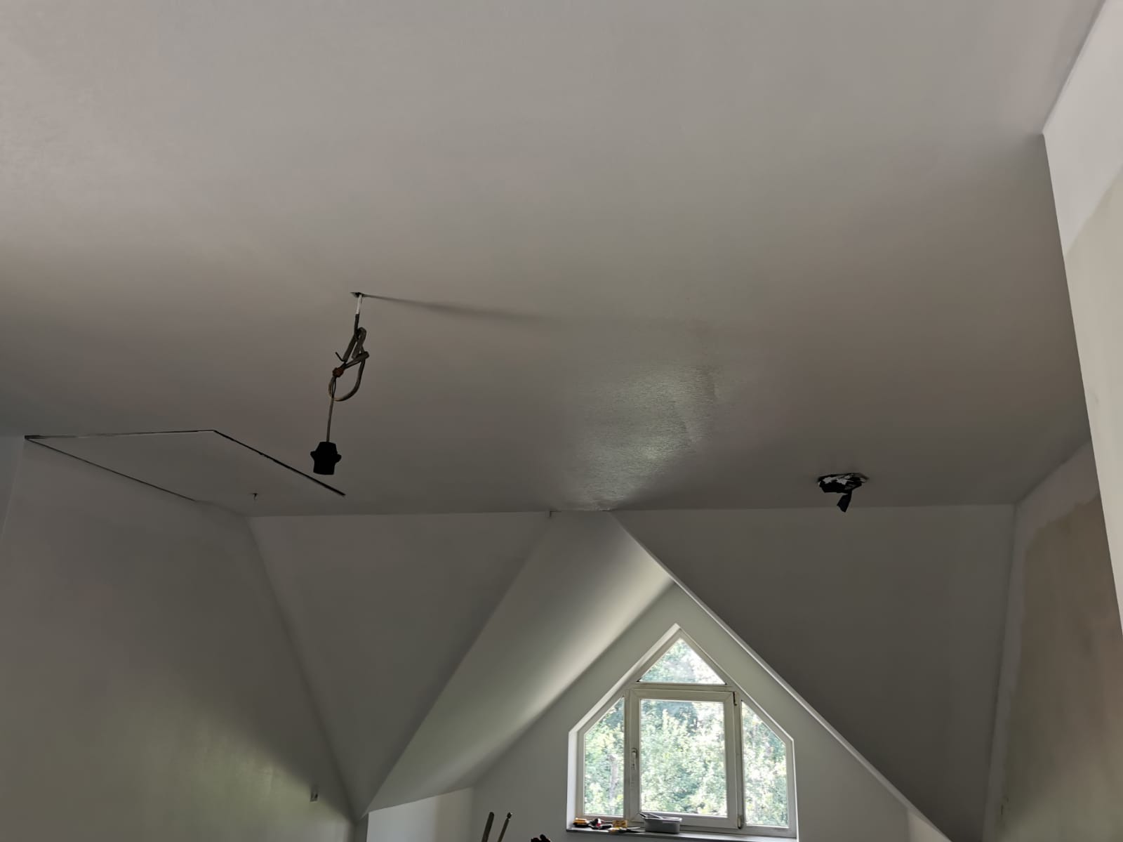 Biały, świeżo pomalowany sufit na poddaszu ze skośnymi ścianami i oknem trójkątnym w tle, widoczne kable i mocowania lamp.