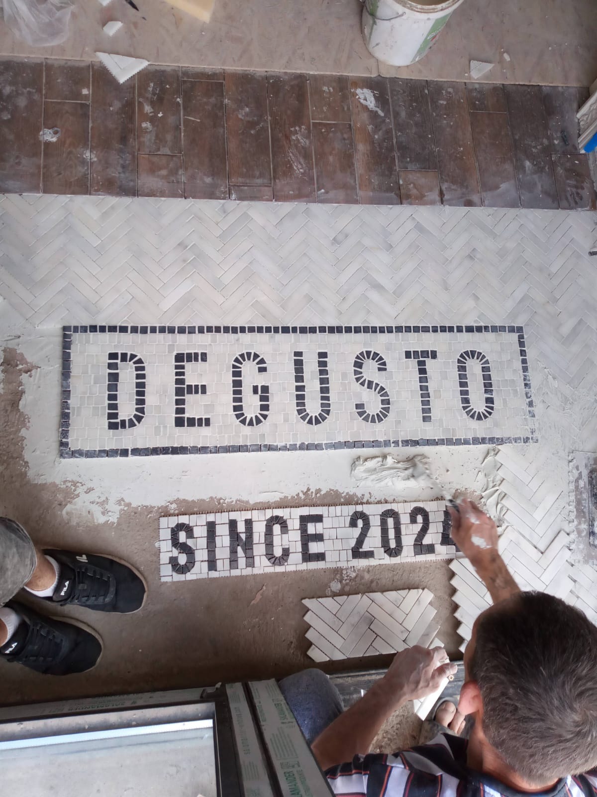 Układanie mozaiki podłogowej z napisem 'DEGUSTO SINCE 2021' w trakcie pracy, widoczne narzędzia i fragmenty nieukończonej powierzchni.