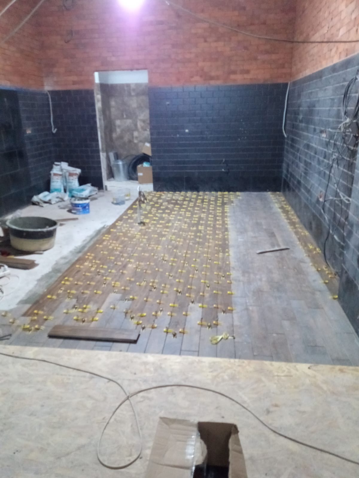 Układanie płytek imitujących drewno na podłodze, widoczne krzyżyki dystansowe, częściowo wykończona ściana z ciemnej cegły, worki z zaprawą w tle.