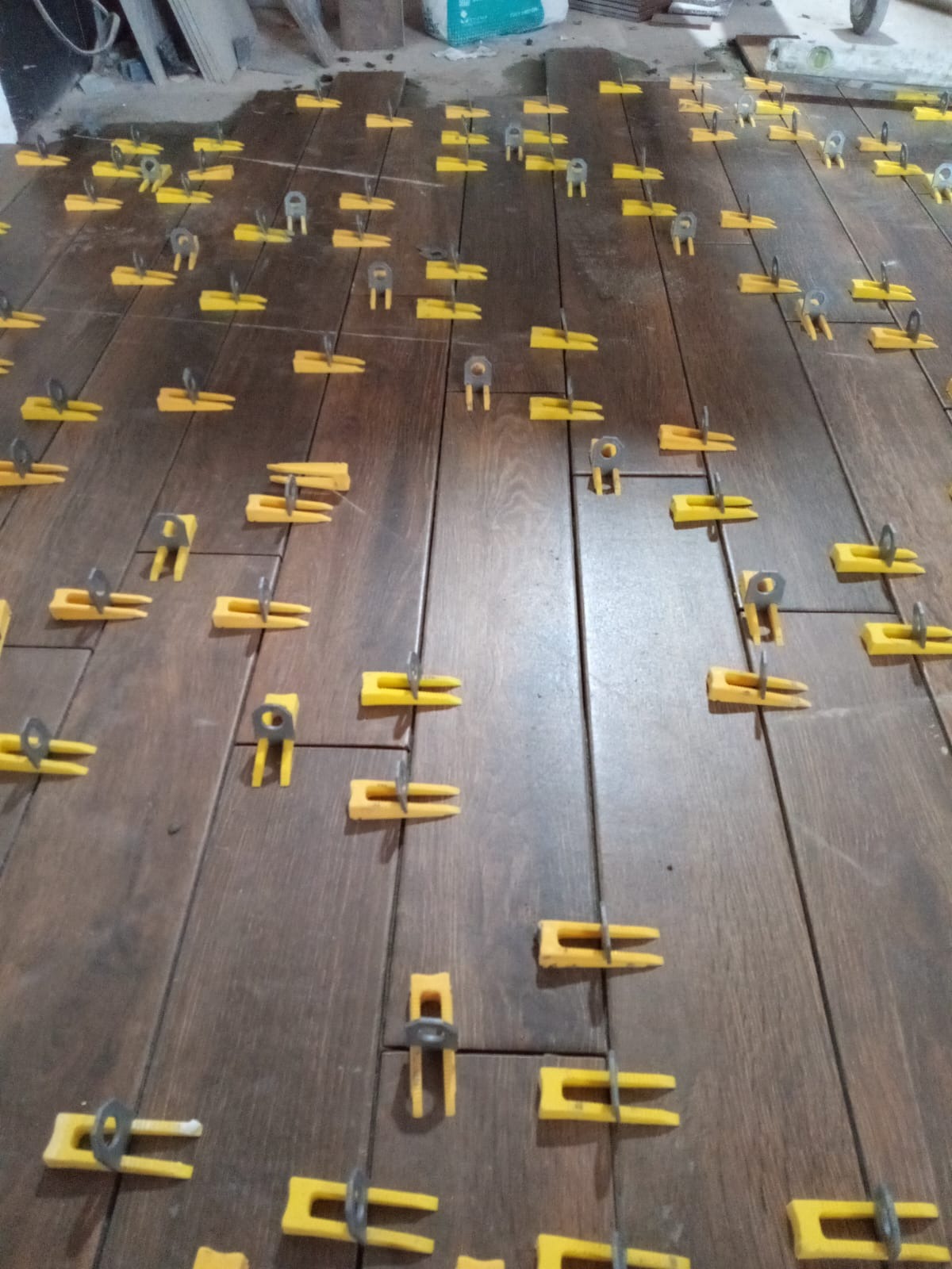 Układanie ciemnobrązowych płytek podłogowych z użyciem żółtych klinów i metalowych pierścieni poziomujących, widoczny worek z zaprawą klejową w tle.