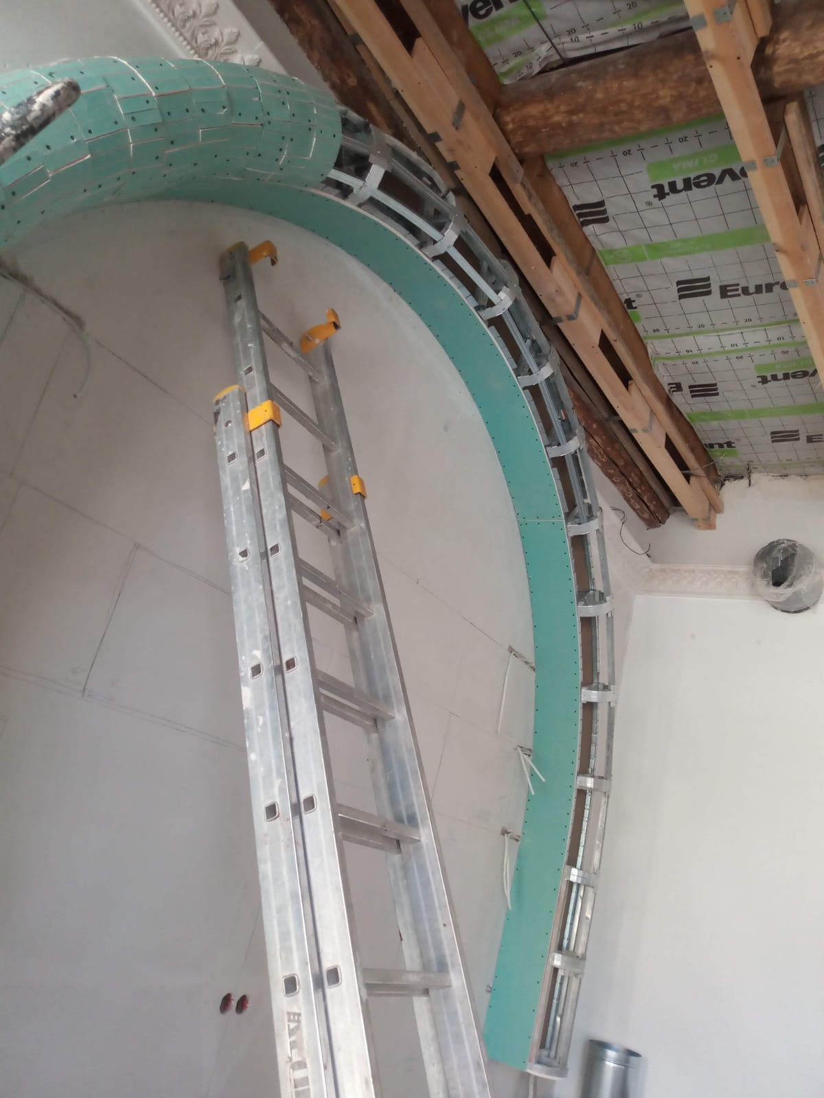 Montaż łuku z płyty karton gips na metalowym stelażu, wsparty drabiną aluminiową, widoczne elementy drewnianej konstrukcji stropu i instalacji wentylacyjnej.