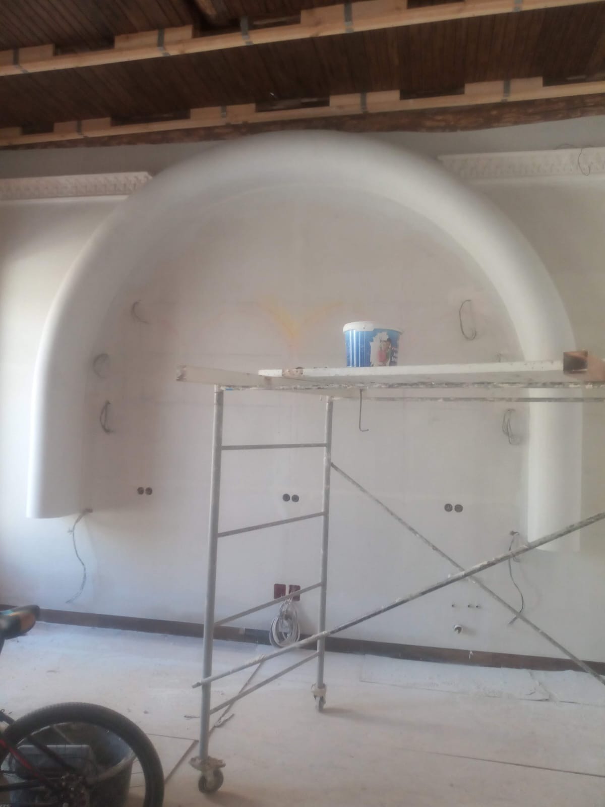 Ściana z wnęką w kształcie łuku w trakcie wykańczania, widoczne przewody elektryczne, puszki pod gniazdka, wiadro z farbą na rusztowaniu, surowy drewniany sufit.