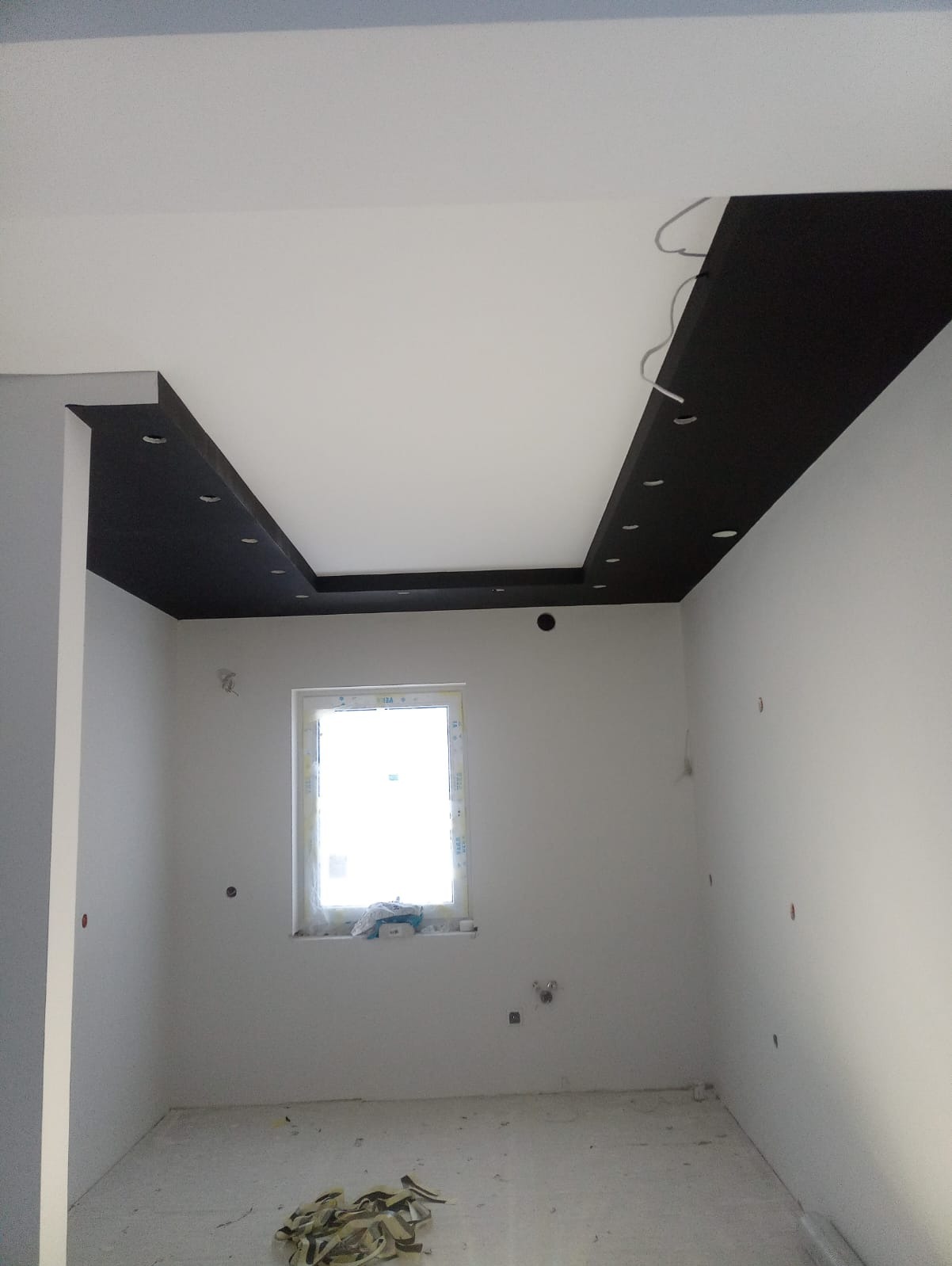 Pokój w trakcie prac wykończeniowych, z podwieszanym sufitem z czarnymi wstawkami i punktowym oświetleniem, widoczne przewody elektryczne, ściany przygotowane do malowania, okno z widocznym parapetem.