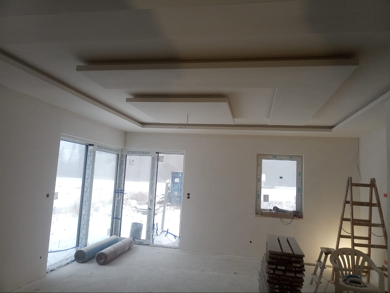 Wnętrze w stanie deweloperskim z zamontowanymi oknami i drzwiami balkonowymi, widoczna konstrukcja sufitu podwieszanego, grzejniki pod folią i drabina oparta o ścianę.