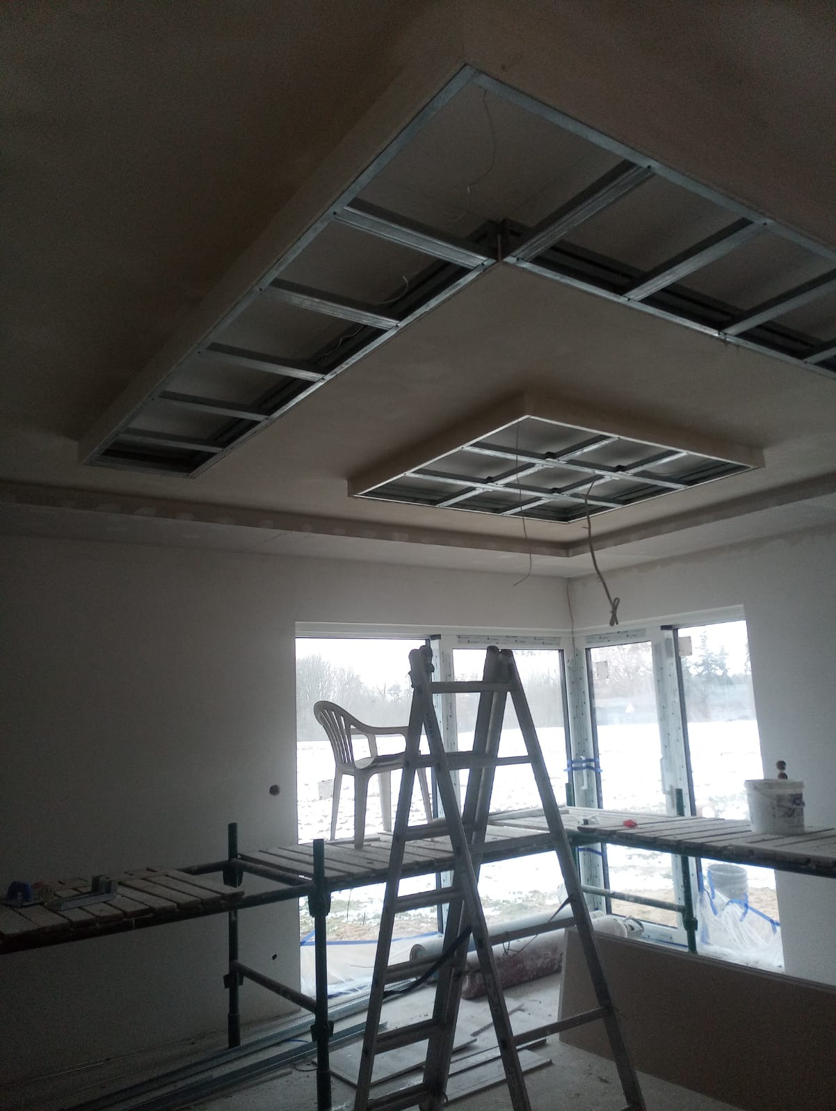Sufit podwieszany z metalową konstrukcją i częściowo zamontowaną płytą gipsowo-kartonową, widok z rusztowania i drabiny w surowym wnętrzu z dużymi oknami.
