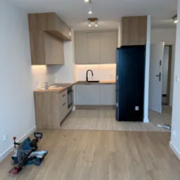 Jasna kuchnia z drewnianymi akcentami, czarną lodówką i narzędziem do cięcia paneli na podłodze. Nowoczesne oświetlenie sufitowe.