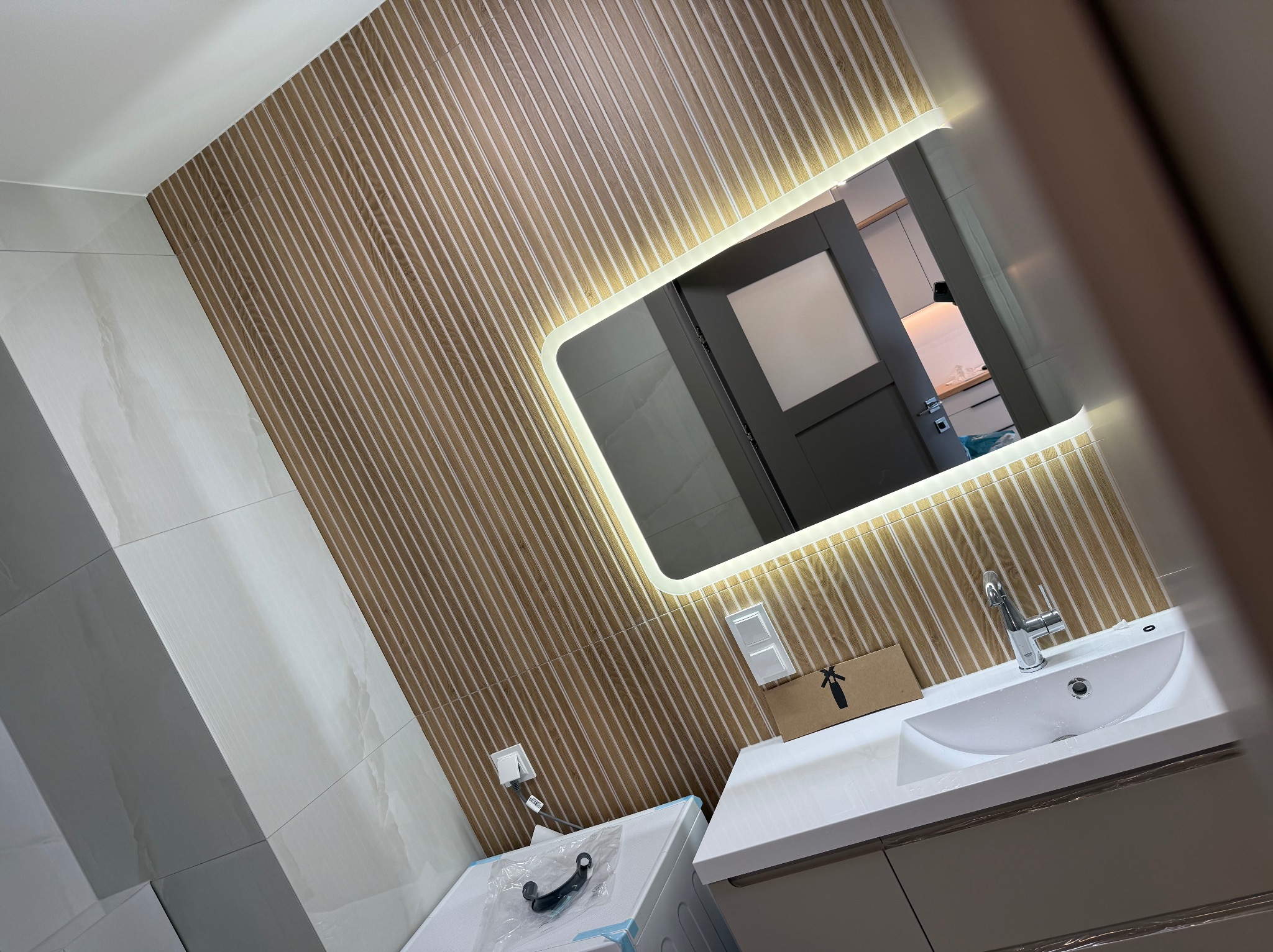 Nowoczesna łazienka z umywalką, podświetlanym lustrem i ścianą w drewniane pasy. W tle widoczne drzwi i fragment kuchni.