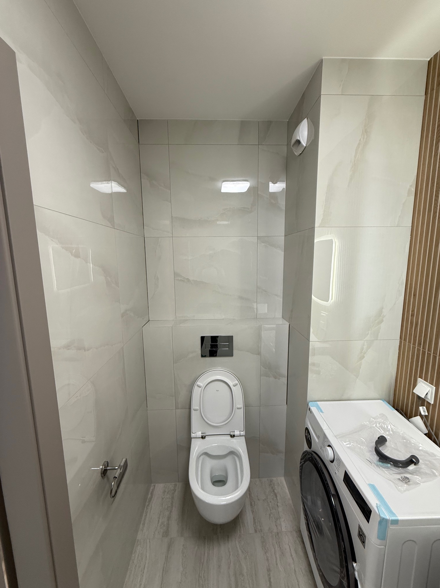 Jasna łazienka po remoncie: białe płytki imitujące marmur na ścianach, podwieszana toaleta z otwartą klapą oraz nowa pralka. Minimalistyczny design.