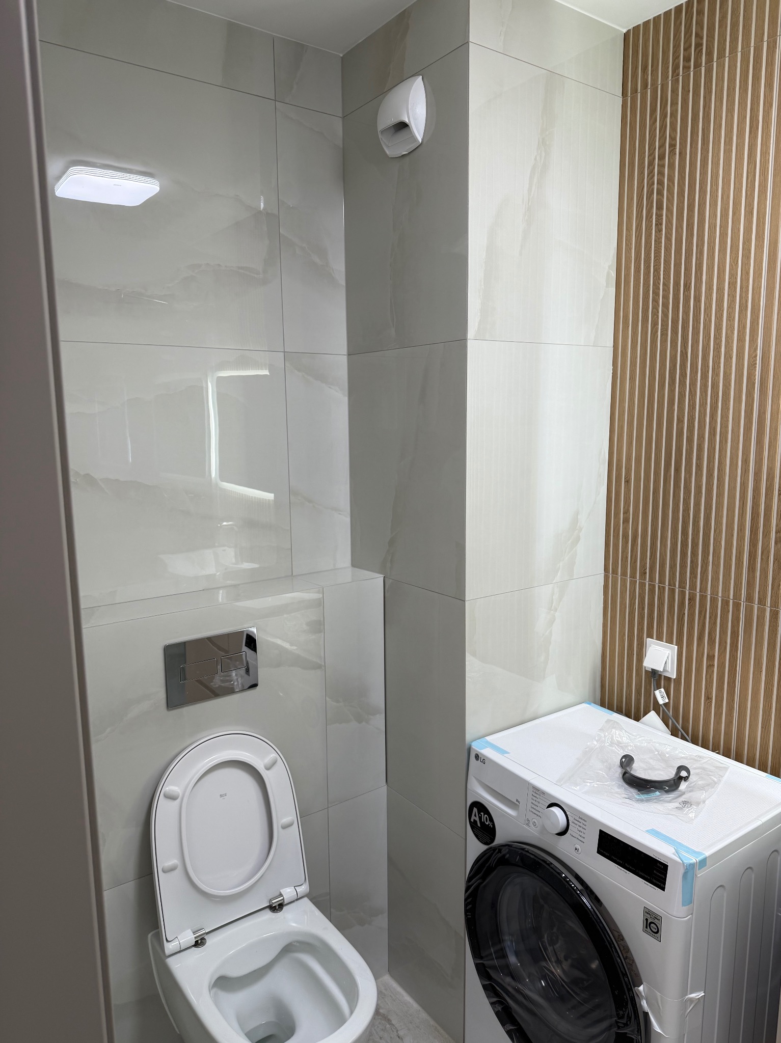 Wykończona łazienka w jasnych płytkach z drewnianym panelem. Widoczna toaleta z podtynkową spłuczką i pralka LG czekająca na podłączenie.