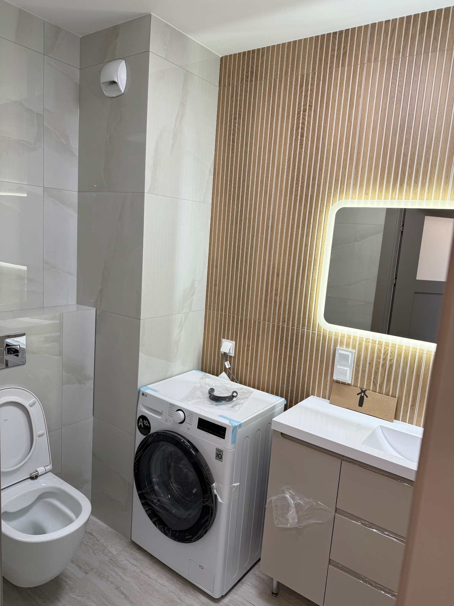 Jasna łazienka z podświetlanym lustrem, drewnopodobnymi panelami ściennymi i białą armaturą. Umywalka z szafką i pralka obok toalety. Nowoczesny design.