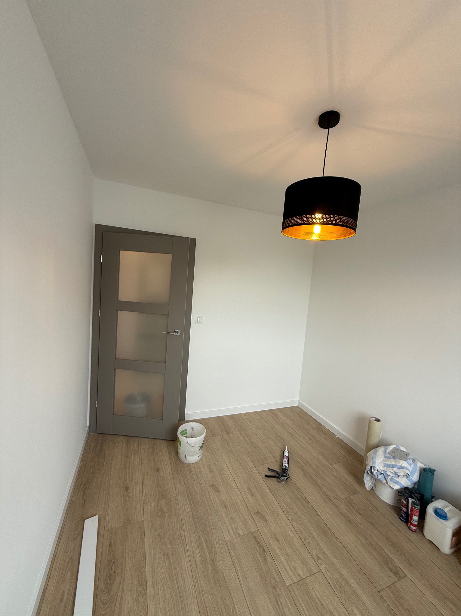 Jasne wnętrze po malowaniu ścian na biało, z drewnianą podłogą, szarą stolarką drzwiową i czarną lampą. Widoczne narzędzia malarskie i wiadro farby.