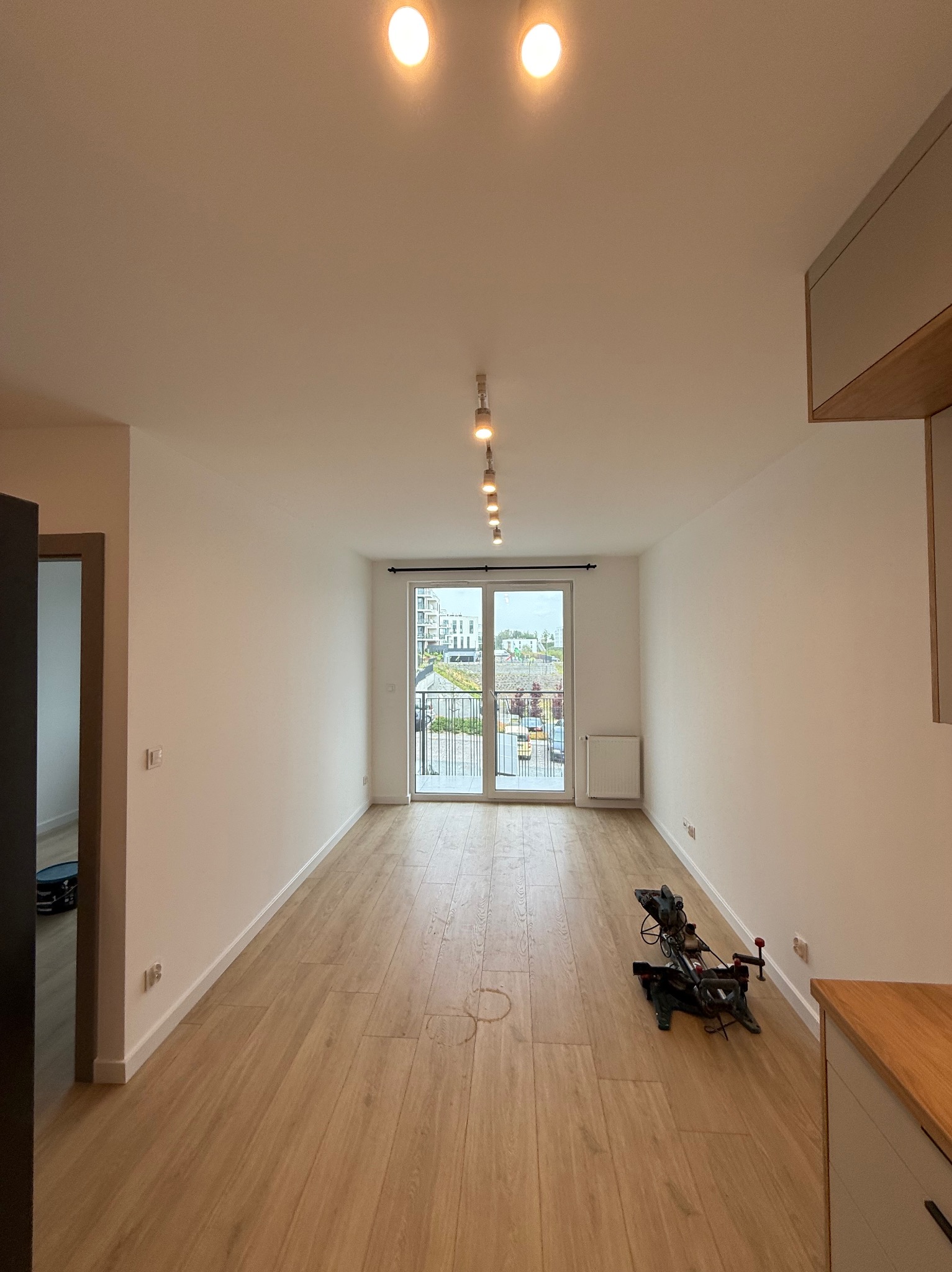 Jasne wnętrze po remoncie: parkiet, białe ściany, widok na balkon. Na podłodze piła ukośnica, sugerująca prace wykończeniowe. Nowoczesne oświetlenie punktowe.