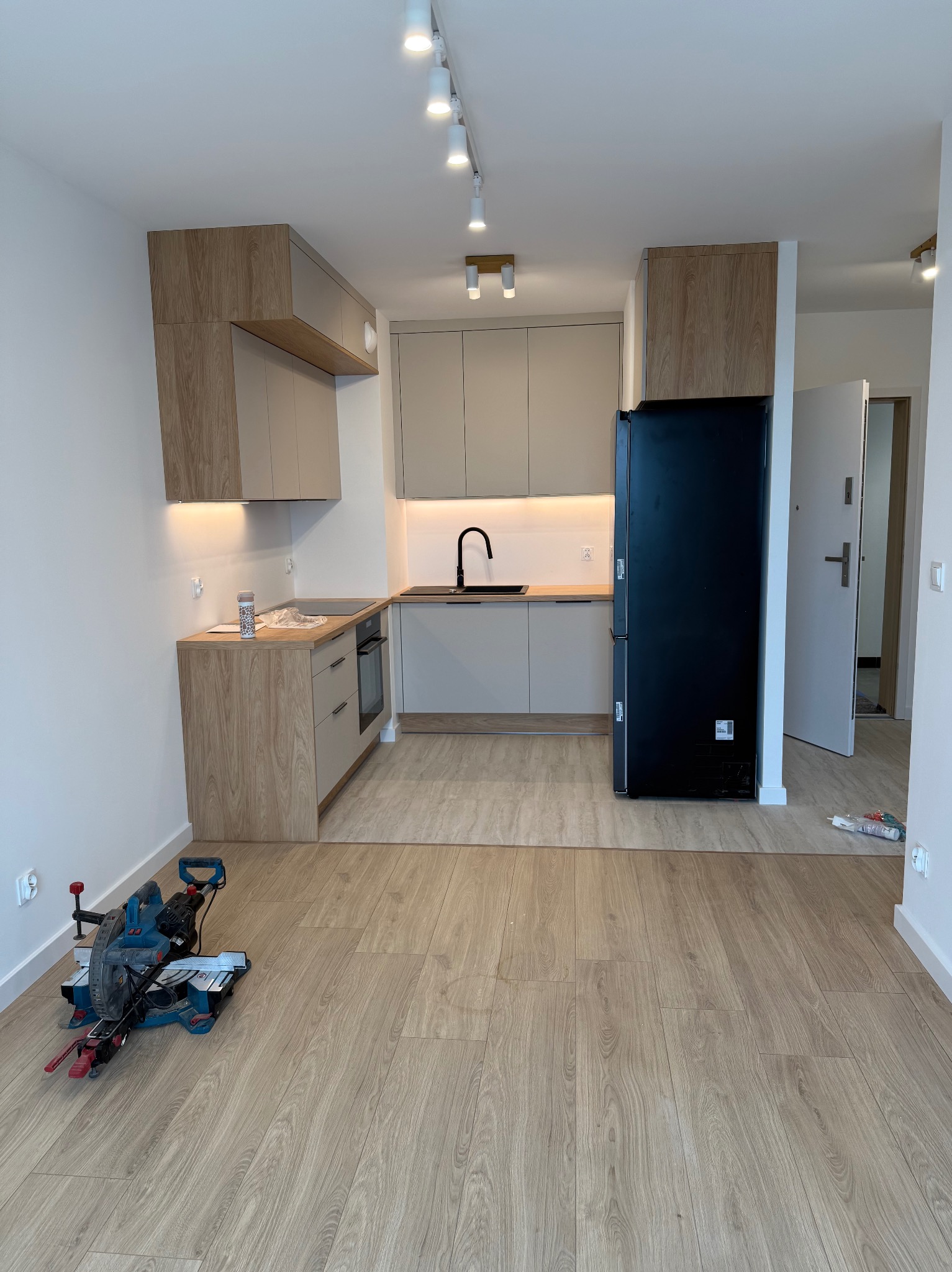 Jasna kuchnia z drewnianymi akcentami, czarną lodówką i narzędziem do cięcia paneli na podłodze. Nowoczesne oświetlenie sufitowe.