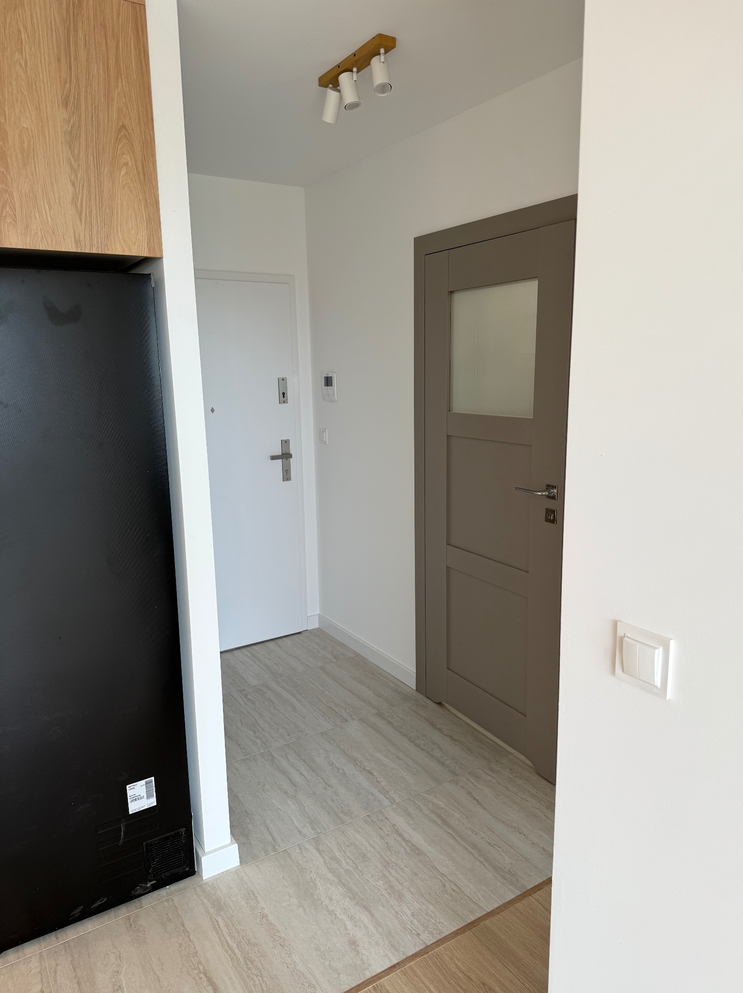 Wykończony przedpokój z beżową podłogą imitującą drewno, dwoje drzwi (białe wejściowe i szare wewnętrzne z szybą), oświetlenie sufitowe i fragment szafy w zabudowie.