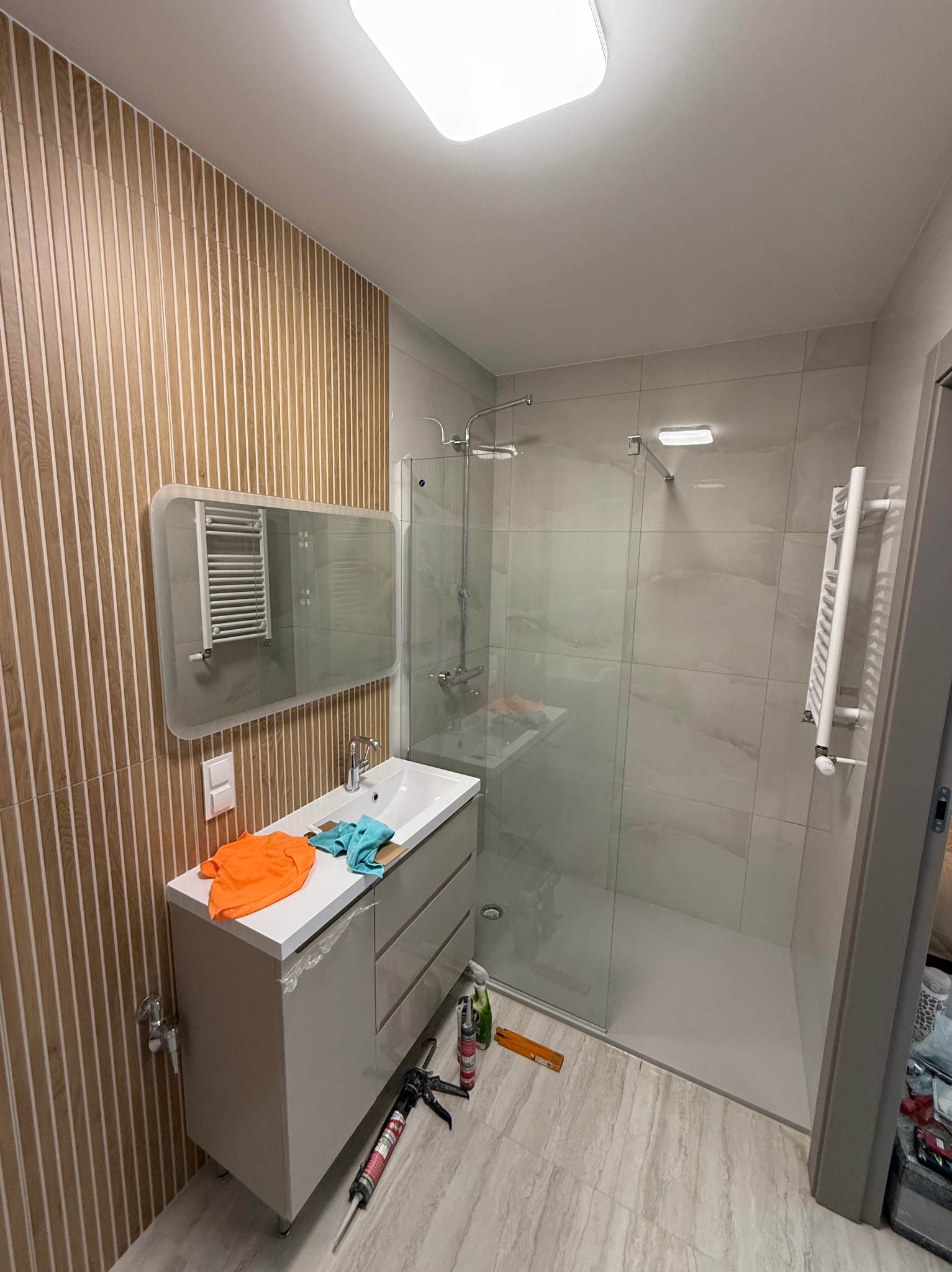 Nowoczesna łazienka po remoncie: drewnopodobne panele ścienne, szklana kabina prysznicowa, umywalka z szafką i lustro z grzejnikiem. Widoczne narzędzia glazurnicze.