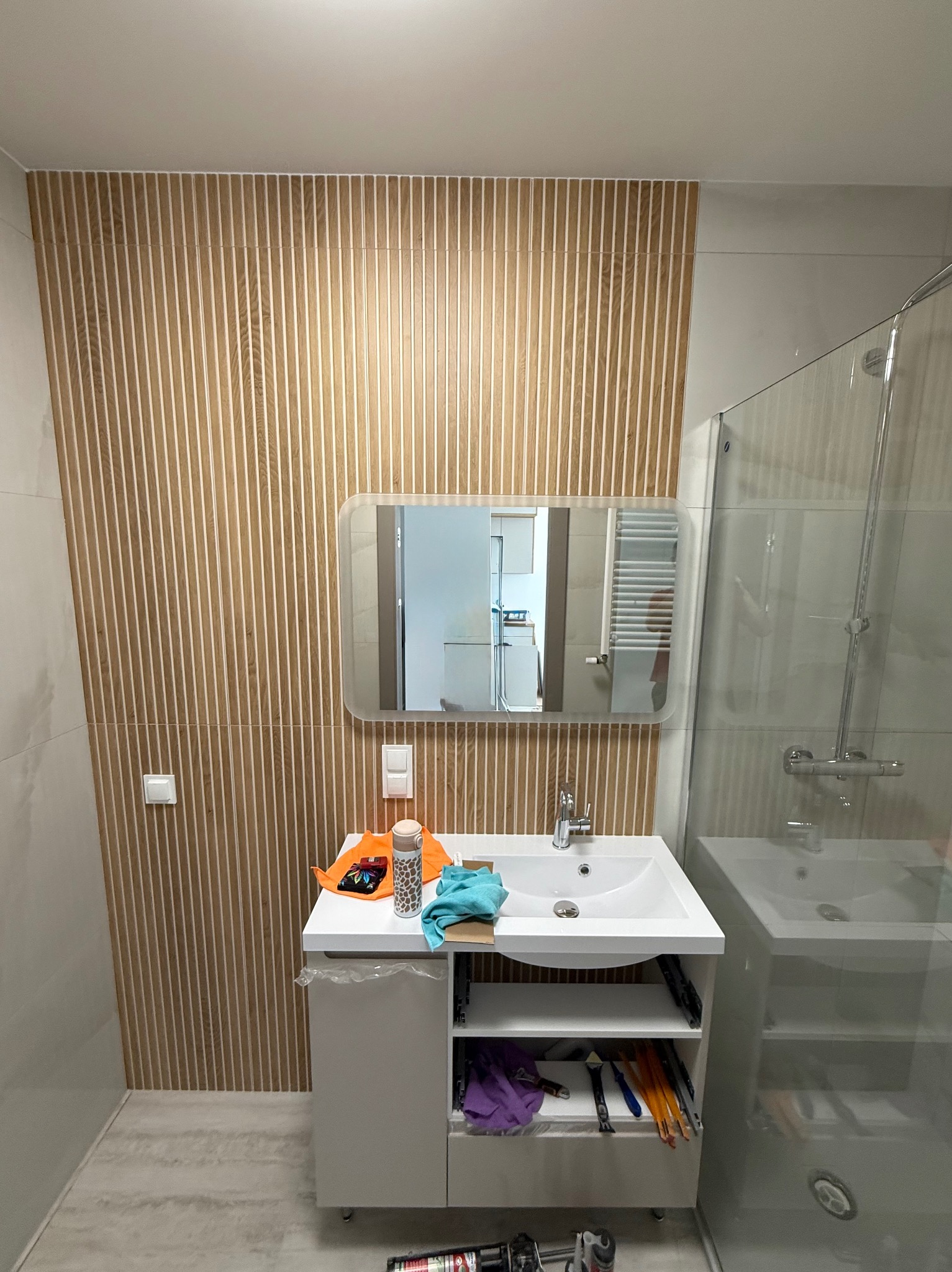 Nowoczesna łazienka po remoncie: drewnopodobne płytki na ścianie, biała szafka z umywalką i lustro. Widoczna kabina prysznicowa i akcesoria.