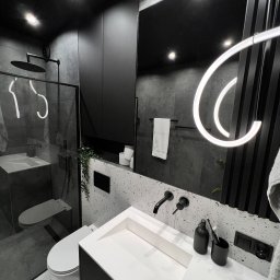 PERFECTBUD GROUP SPÓŁKA Z OGRANICZONĄ ODPOWIEDZIALNOŚCIĄ - Nowoczesna łazienka w czerni i bieli: minimalistyczna umywalka, deszczownica za szklaną ścianką, designerskie oświetlenie i płytki lastryko.