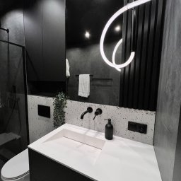 PERFECTBUD GROUP SPÓŁKA Z OGRANICZONĄ ODPOWIEDZIALNOŚCIĄ - Nowoczesna łazienka w czerni i bieli: biała umywalka z czarną armaturą, lustro z dekoracyjnym oświetleniem LED, szare płytki i akcenty roślinne. Minimalistyczny design.