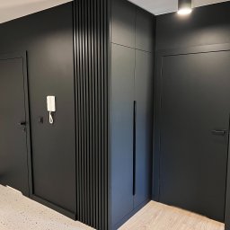 PERFECTBUD GROUP SPÓŁKA Z OGRANICZONĄ ODPOWIEDZIALNOŚCIĄ - Czarne, minimalistyczne wnętrze: zabudowa szafy z pionowymi lamelami, dwie pary drzwi, biały domofon na ścianie. Podłoga w jasnym odcieniu drewna.