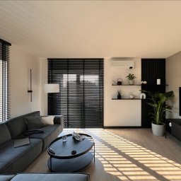 PERFECTBUD GROUP SPÓŁKA Z OGRANICZONĄ ODPOWIEDZIALNOŚCIĄ - Nowoczesny salon z szarym narożnikiem, okrągłym stolikiem i drewnianymi żaluzjami, przez które wpada ciepłe światło słoneczne. Minimalistyczne półki z dekoracjami.